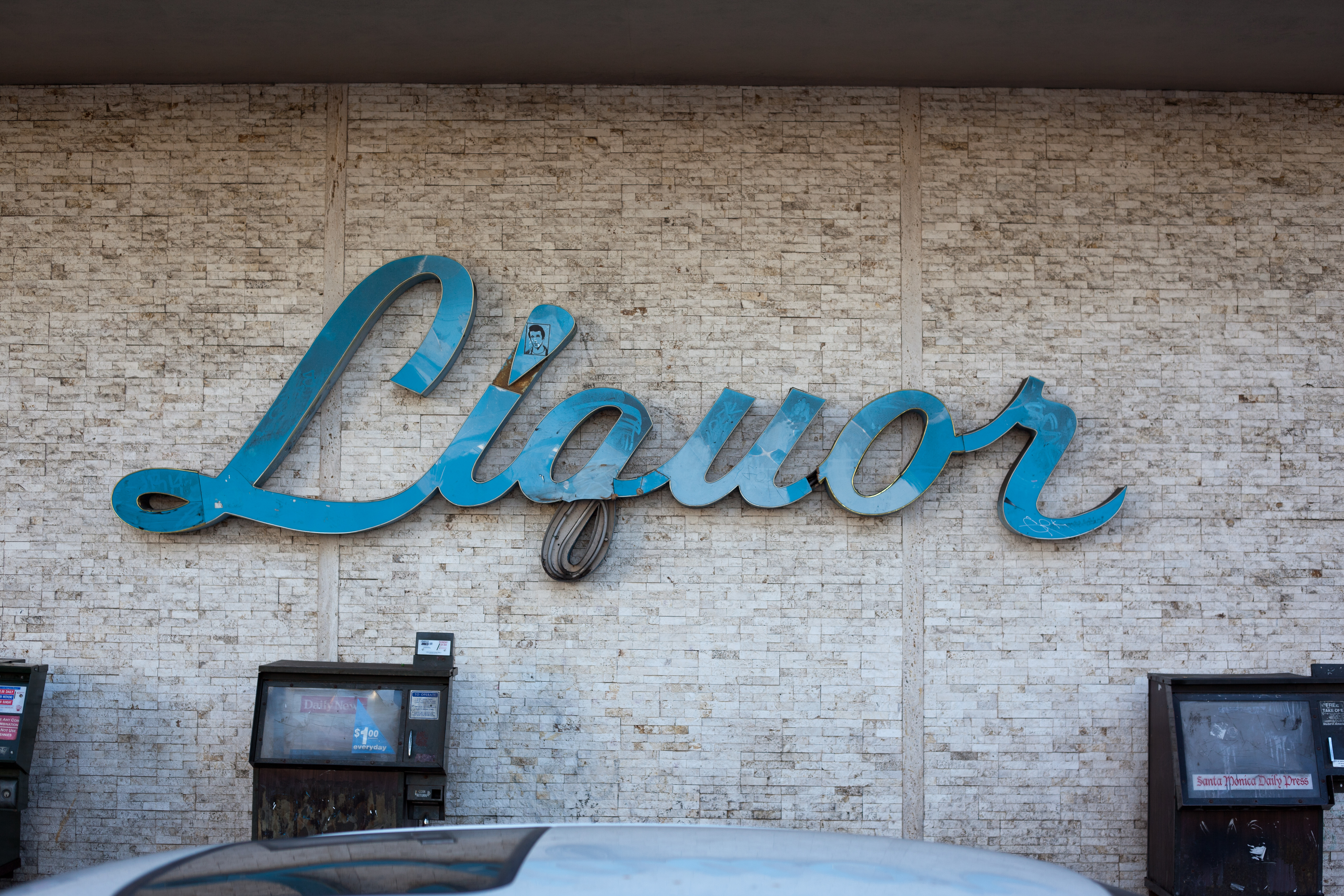 Turquoise neon sign reading 'Liquor' mounted on a textured stone wall above newspaper racks.