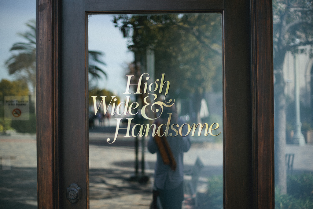 Gold leaf script lettering on a glass storefront door reading 'High Wide & Handsome' reflecting outdoor street scenery in California, shot with a Canon 5D Mark II.