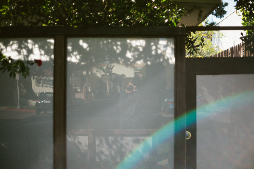 Photographer self-portrait reflected in a frosted glass door holding a Canon DSLR with a vibrant rainbow lens flare across the frame in California, shot with a Canon 5D Mark II.