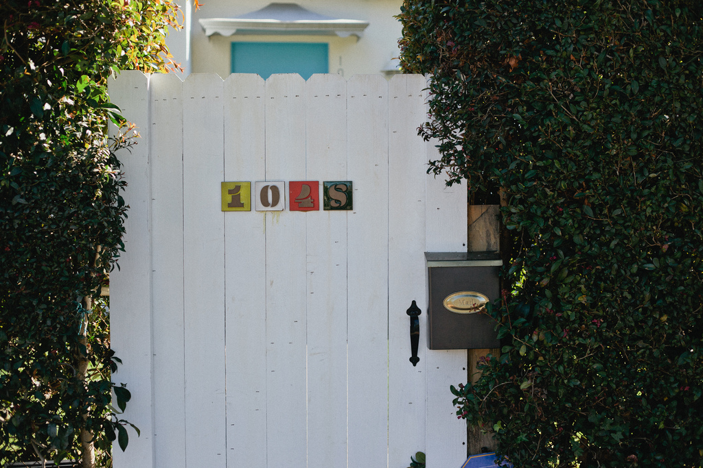 White garden gate framed by hedges, showing house number tiles 1028 and a bronze mailbox.