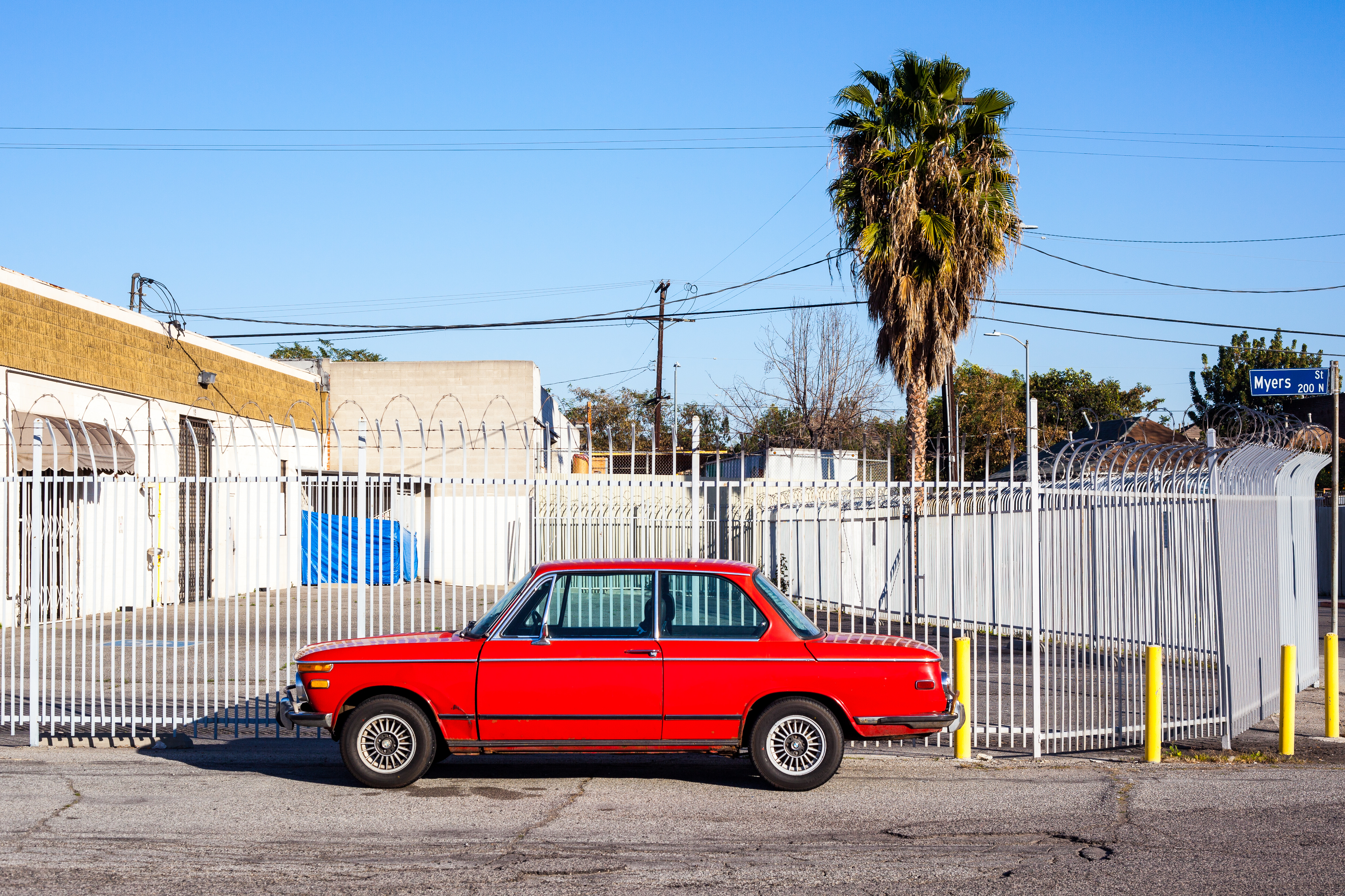 Classic red BMW 2002 coupe parked in front of a white security fence and tall palm tree.