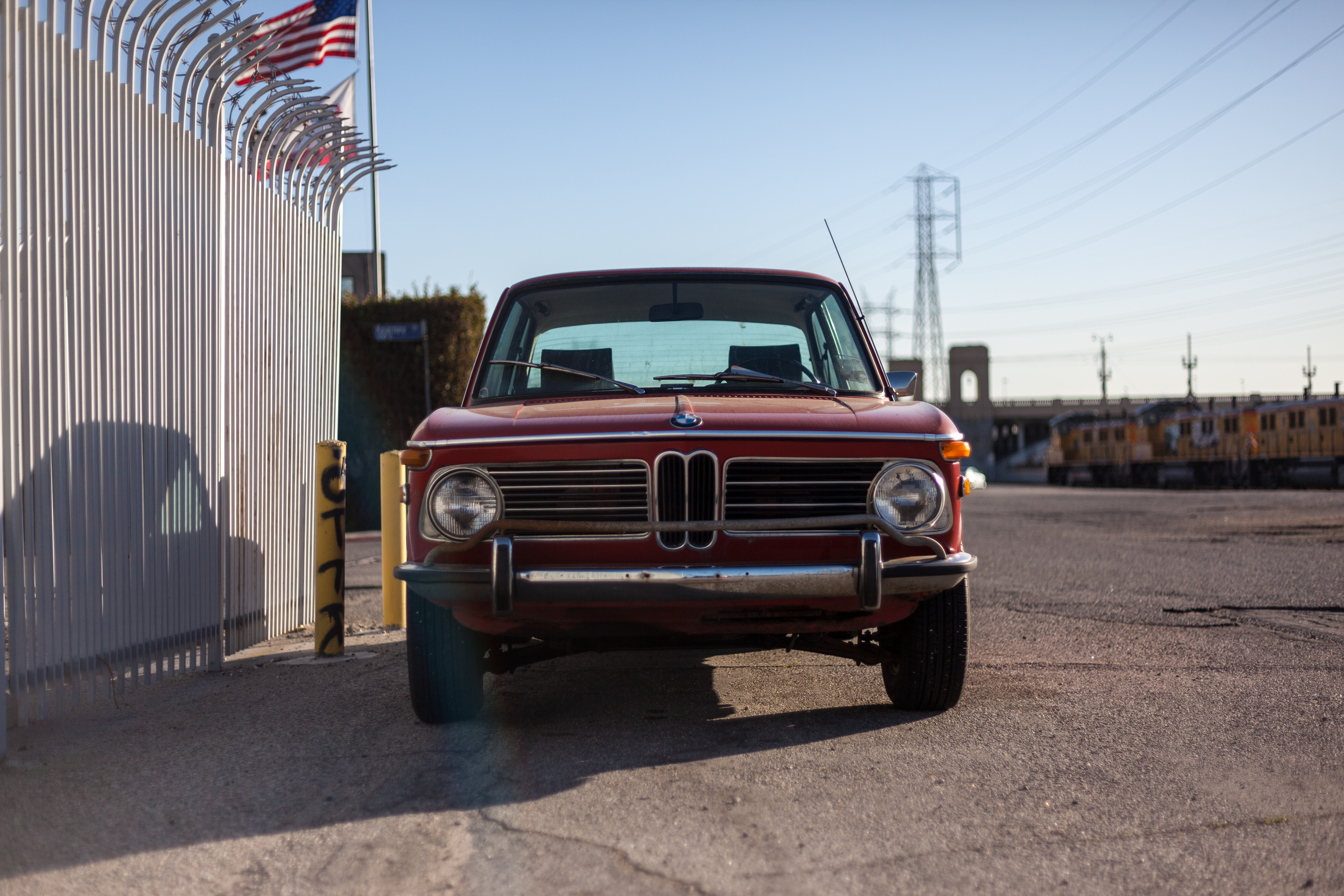 Front view of a vintage red BMW 2002 coupe parked next to a white iron security fence.
