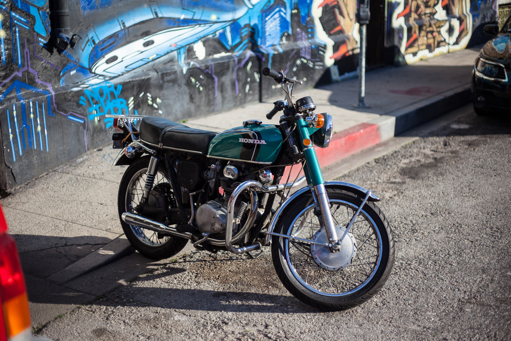 Vintage teal-green Honda CB motorcycle parked curbside against a vibrant graffiti mural wall in California.
