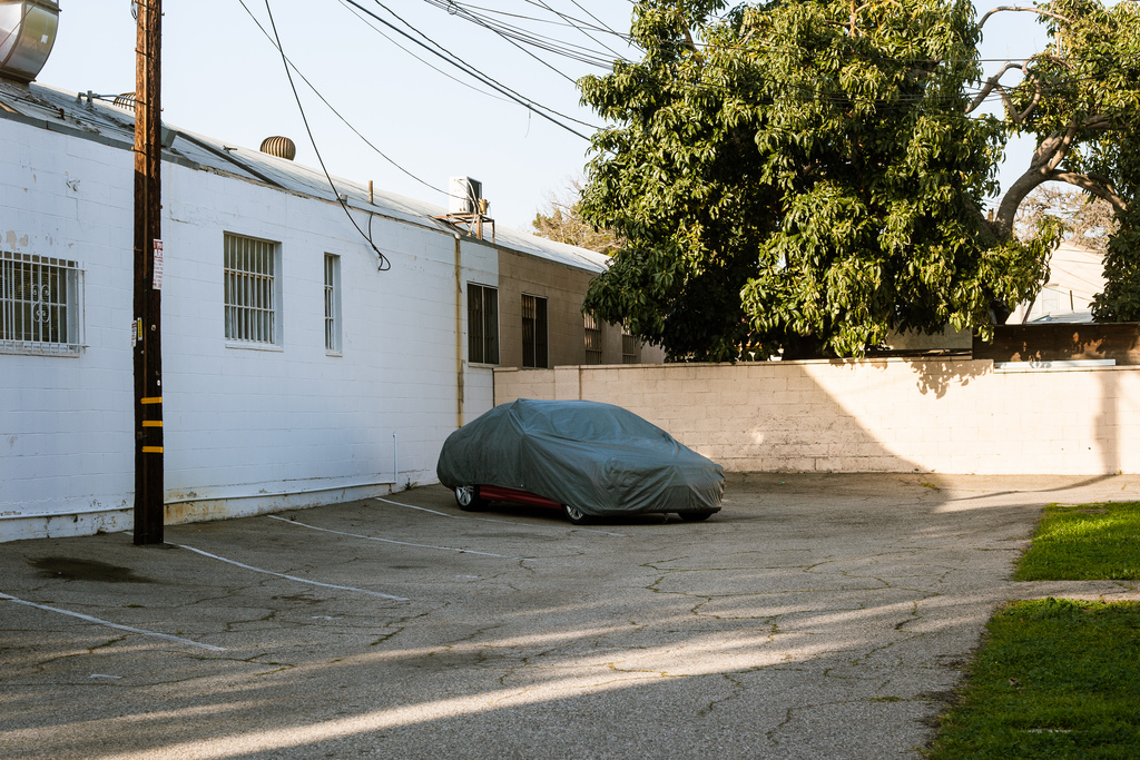 Car covered in a gray protective tarp parked in an asphalt lot beside a white building.