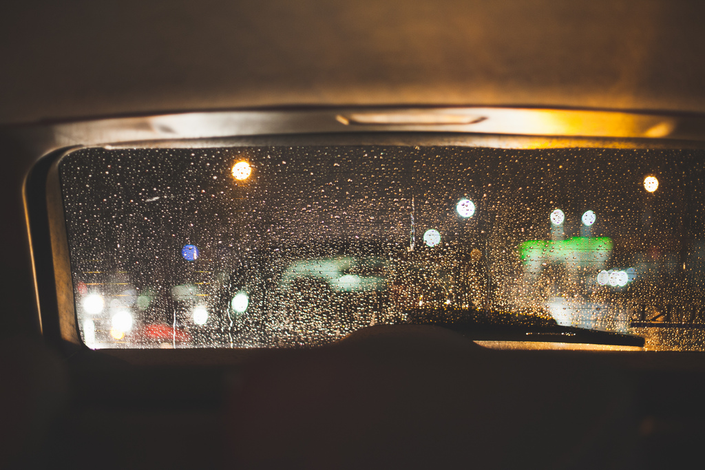 View looking out the rain-streaked rear window of a car at night with glowing amber and green streetlights blurred through water droplets in California, shot with a Canon 5D Mark II.
