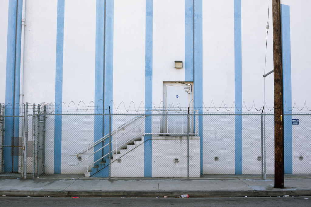 White industrial building with blue stripes, an exterior metal staircase, and barbed wire.