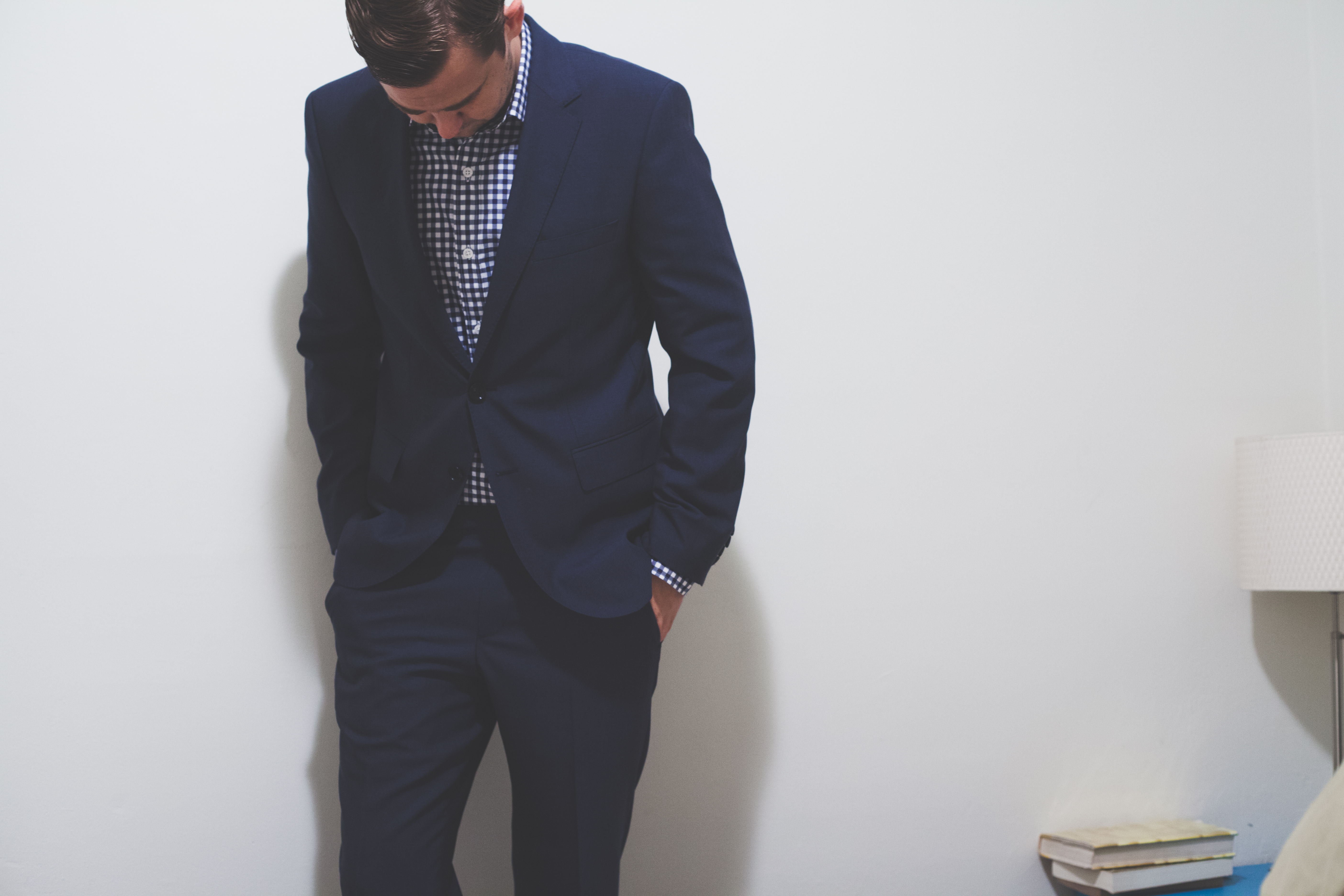 Man in a navy suit and gingham check shirt standing against a white wall looking downward.