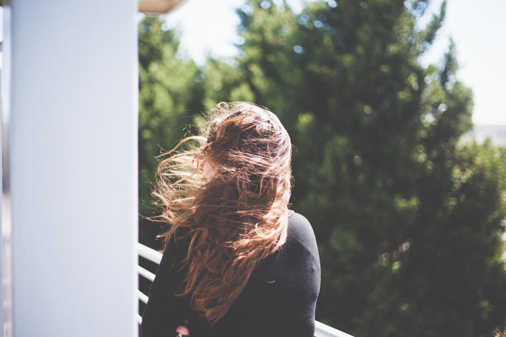 Maggie with windblown long brown hair standing outdoors by a white balcony railing.