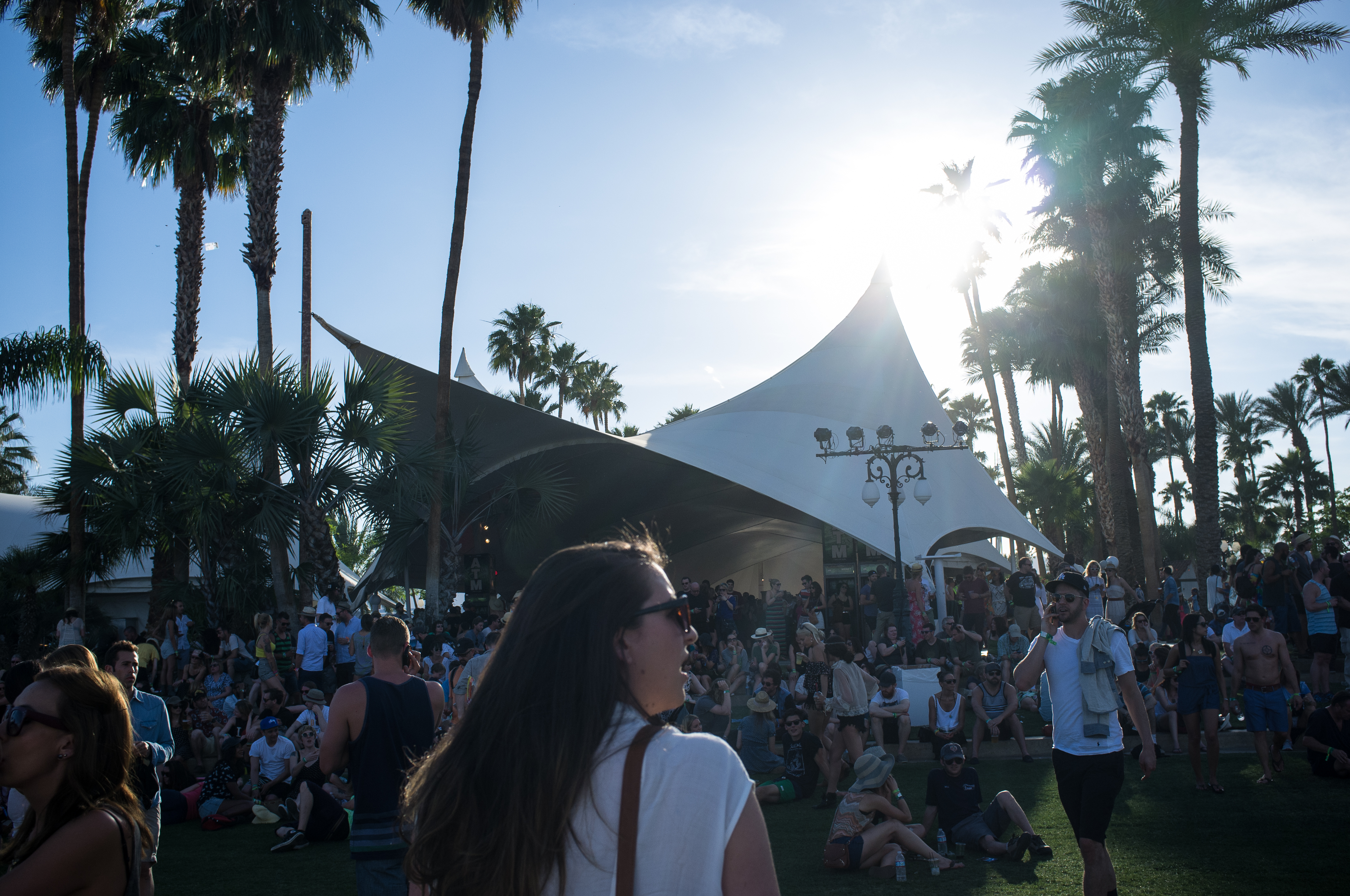 Festivalgoers relaxing in palm tree shade near a white peaked canopy pavilion at Coachella.