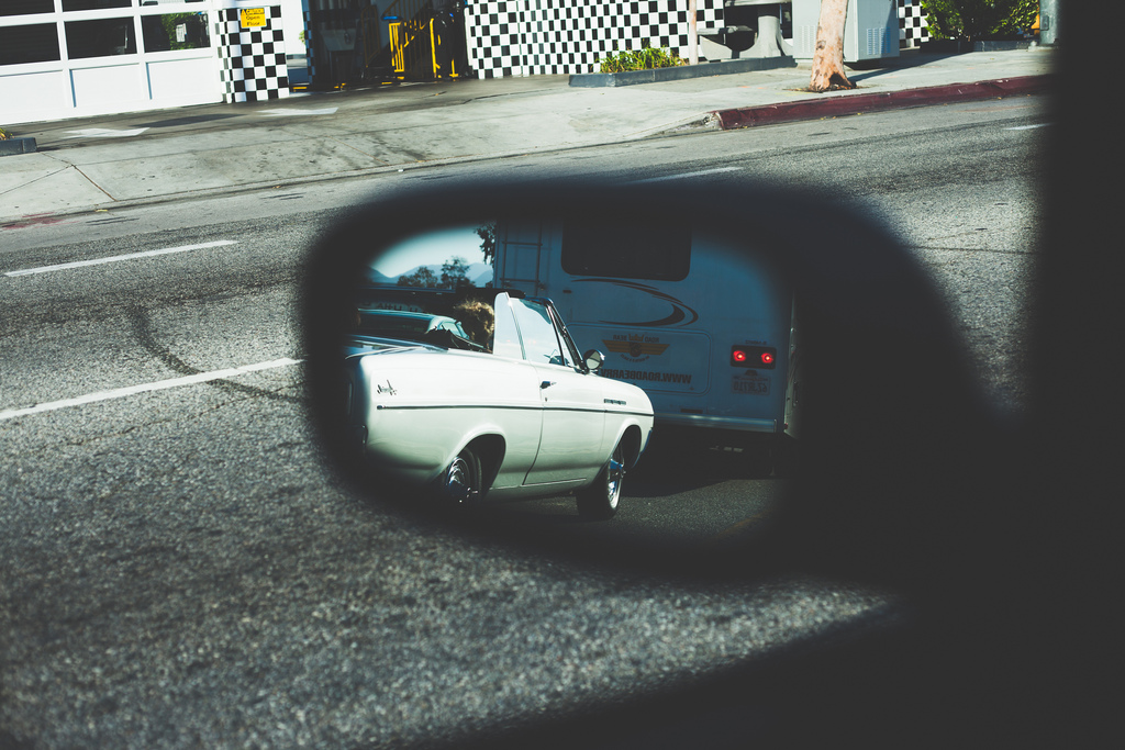 Car side mirror reflection showing a classic white convertible following on a city street beside a checkered building wall, shot with a Canon 5D Mark II.