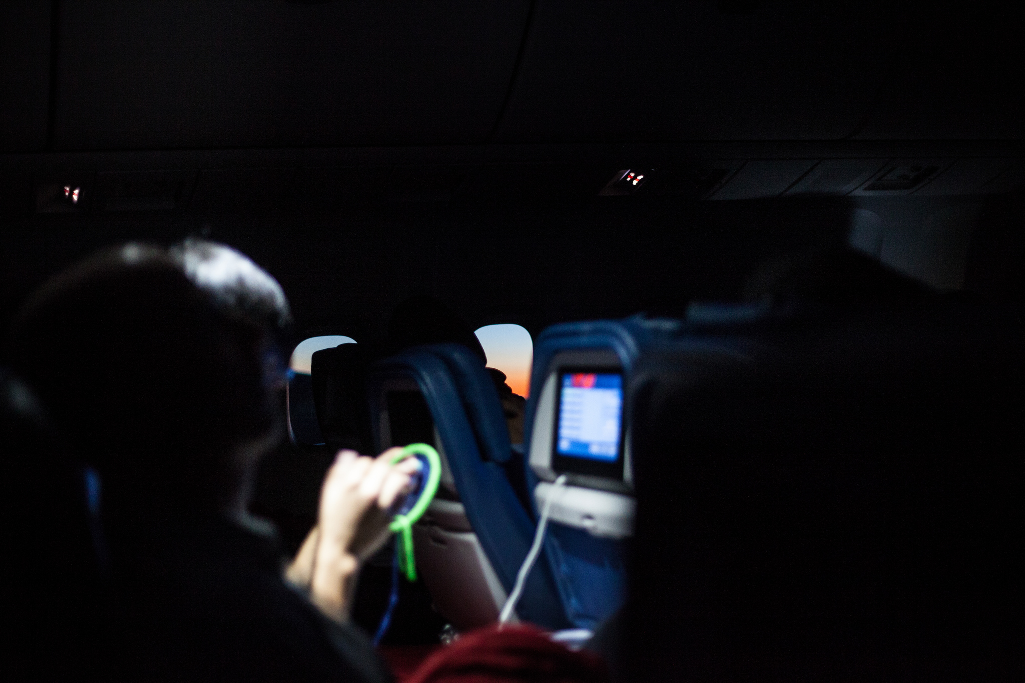 Darkened airplane cabin during flight with glowing screens and an oval window showing twilight.