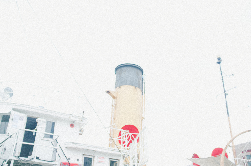 Low-angle view of a vintage steamboat's cream and red smokestack against a bright white sky in New York City.