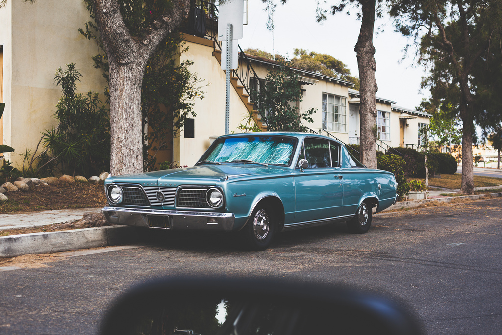 Vintage turquoise Plymouth Barracuda fastback parked curbside before a yellow apartment building.