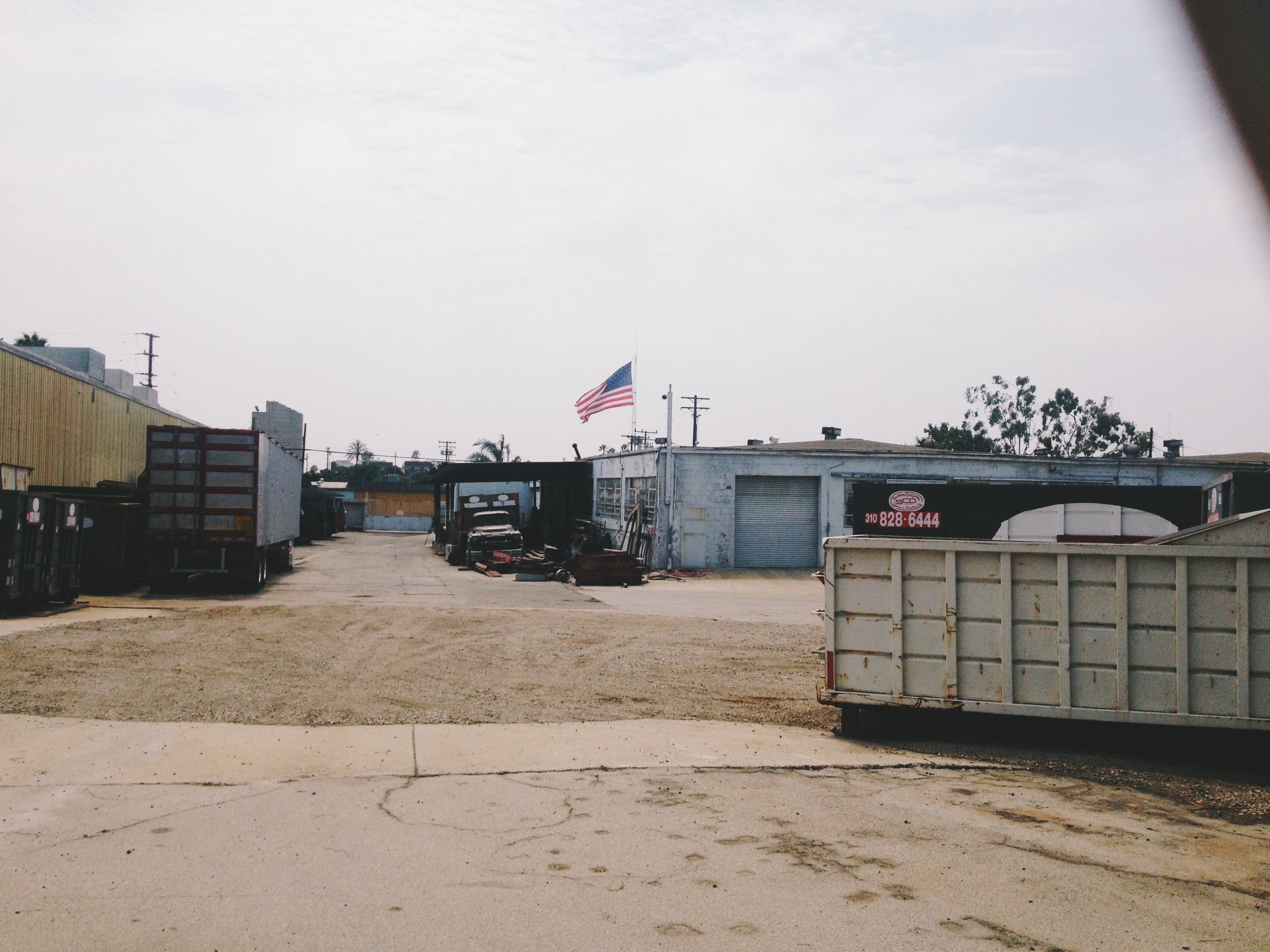 Industrial warehouse yard with an American flag flying high and a large dumpster in the foreground.