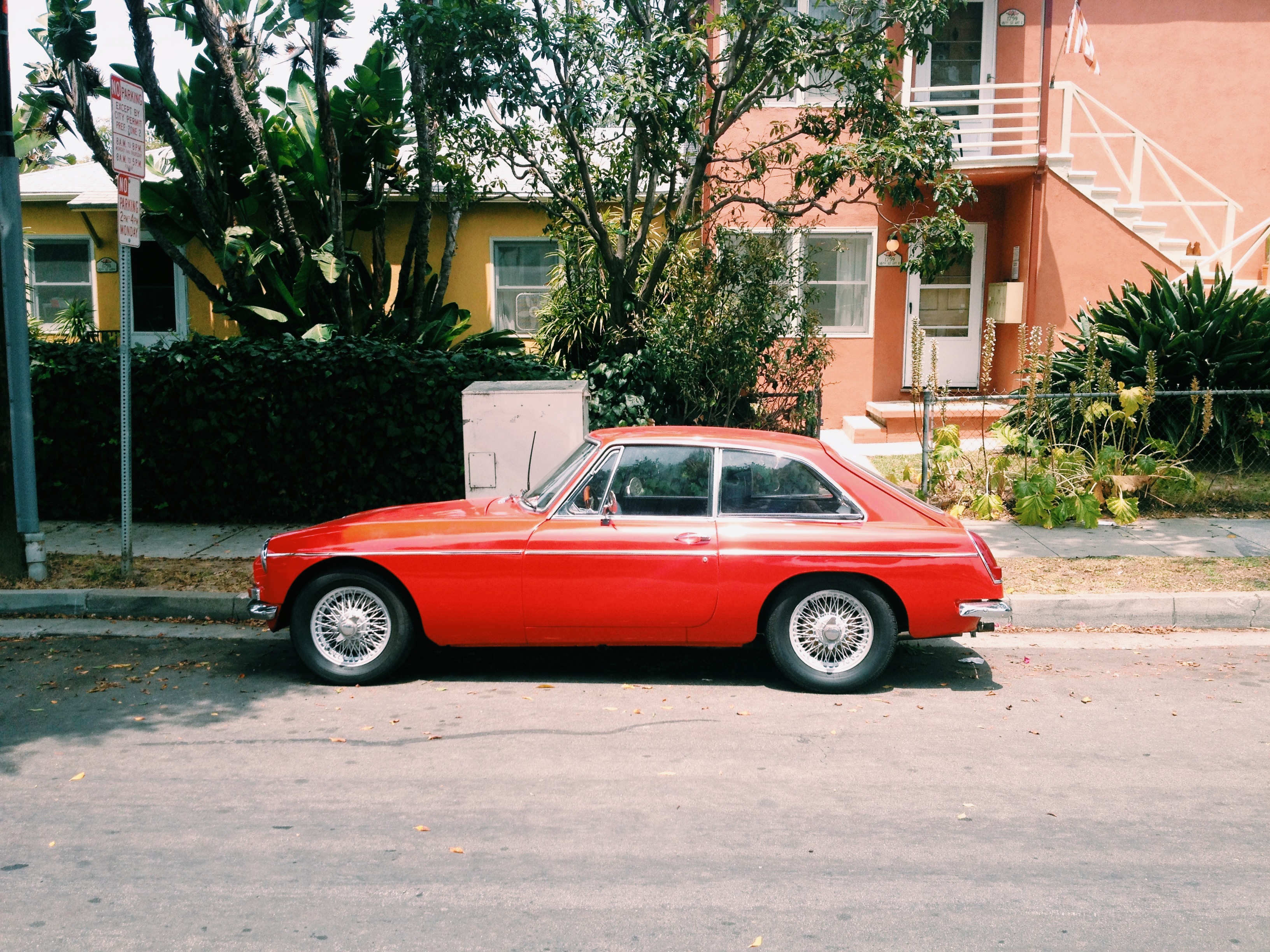 Red vintage MGB GT sports coupe parked curbside in front of a peach apartment house.