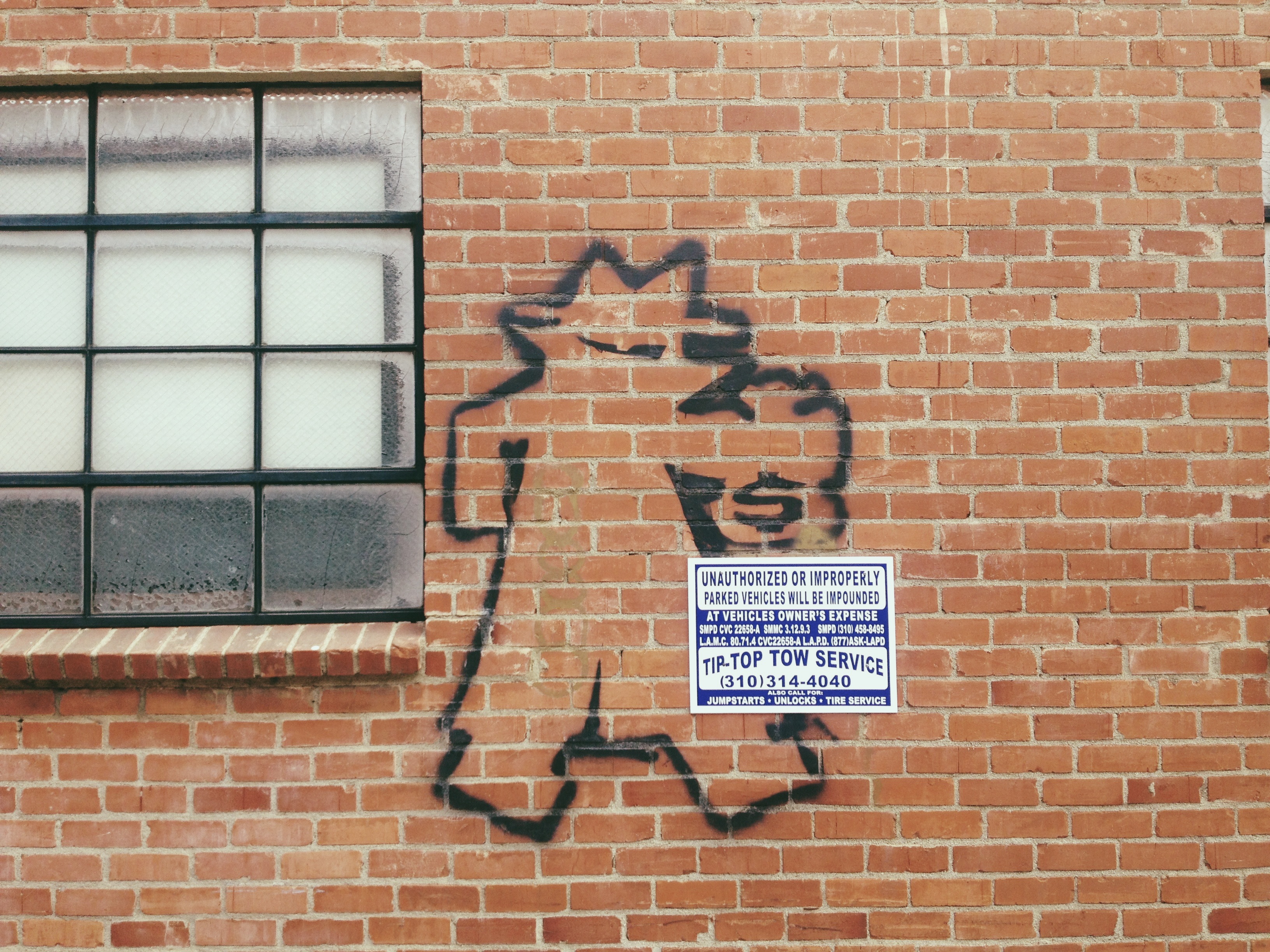 Red brick exterior wall with black spray paint graffiti next to a blue 'TIP-TOP TOW SERVICE' parking sign.