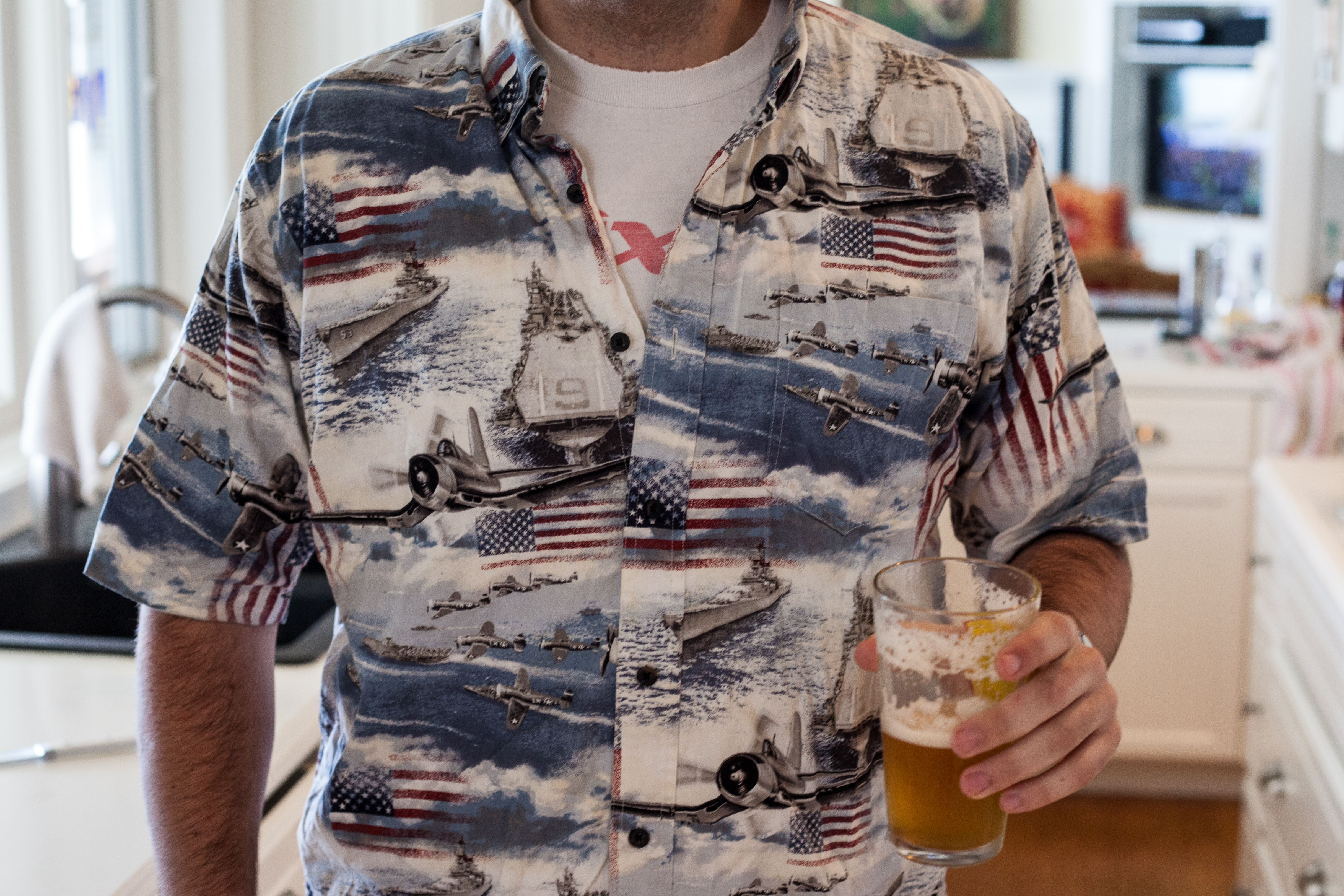 Man in a patriotic flag-patterned button-down shirt holding a beer in a kitchen.