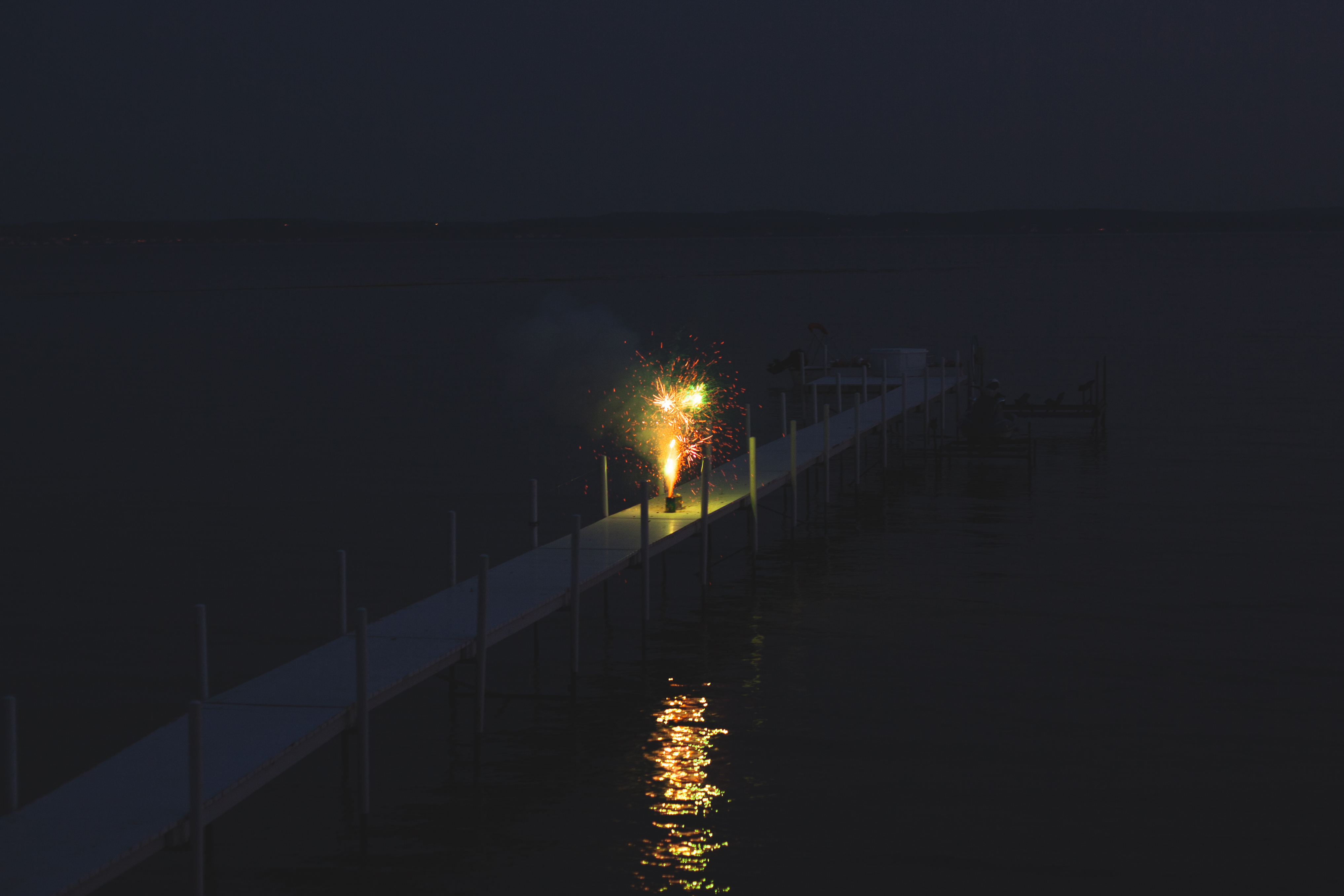 Wooden dock on Lake Michigan with a fountain firework sparkling green and gold over the water at night.