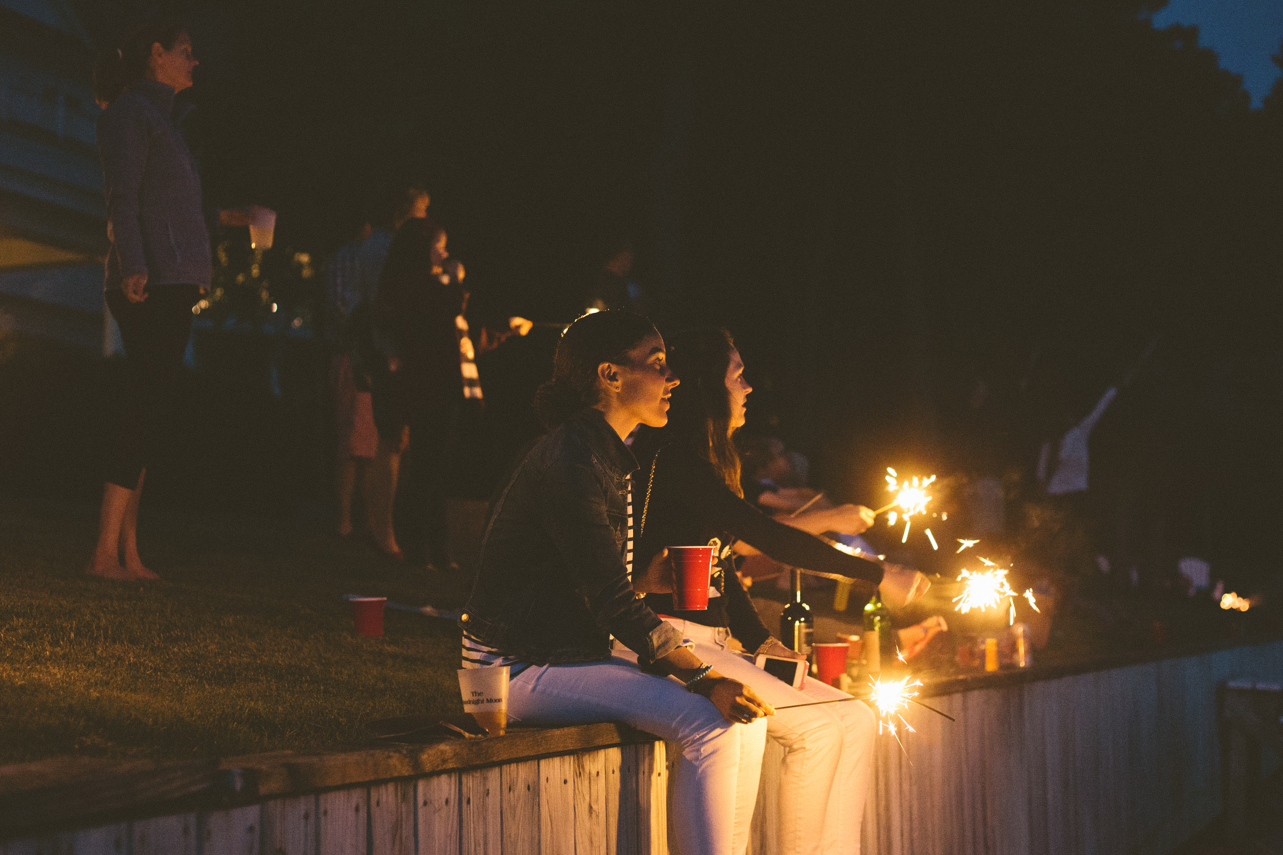 Group of friends sitting along a wooden bulkhead at night holding glowing sparklers and red plastic party cups celebrating in Harbor Springs, shot with a Canon 5D Mark II.