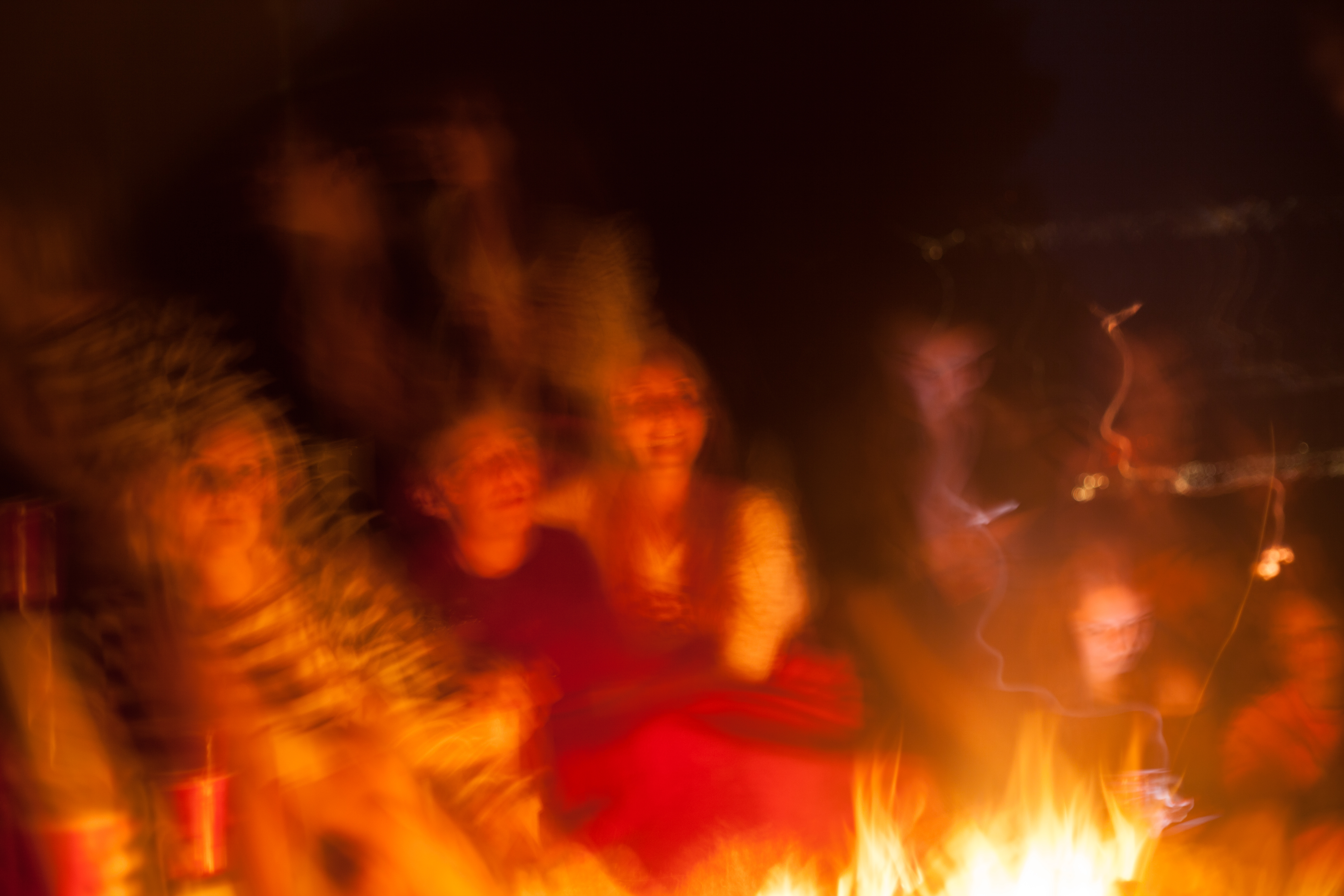 Warm, dreamy motion-blurred photograph of people gathered around the leaping orange flames of a campfire at night, shot with a Canon 5D Mark II.
