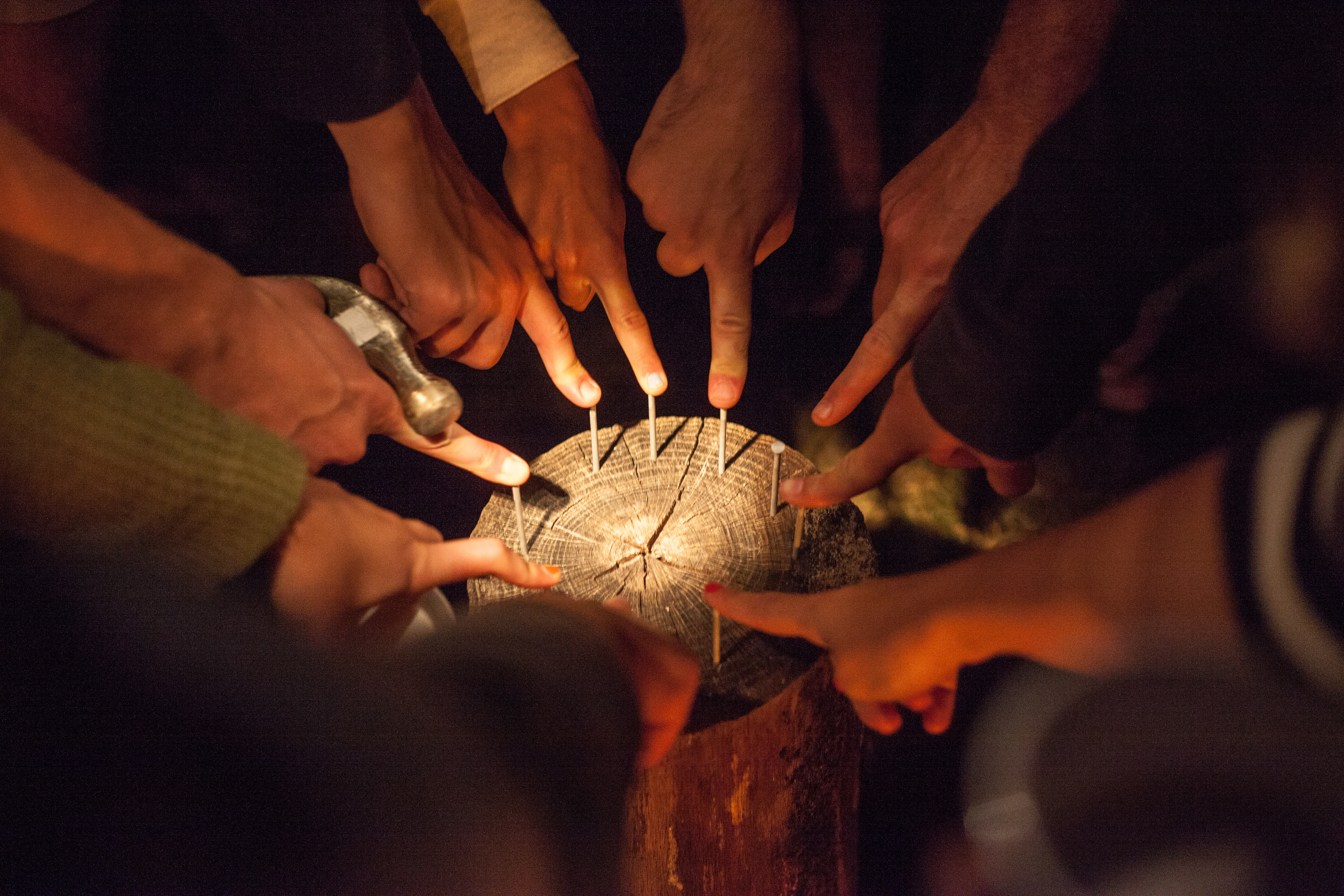 Close-up overhead view of hands pointing down at nails arranged in the end grain of a tree log during a game of Hammerschlagen around a fire, shot with a Canon 5D Mark II.