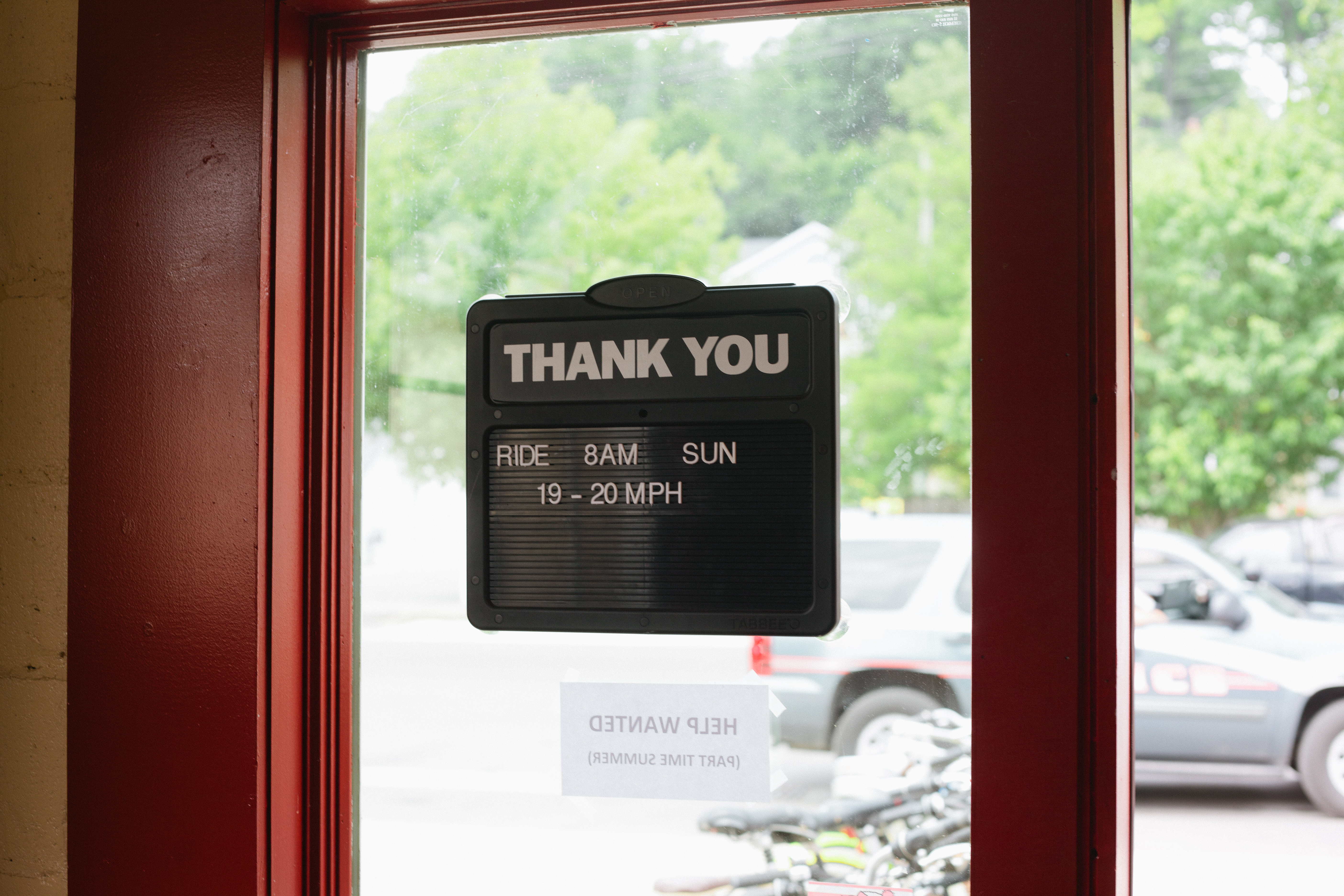 Sign reading 'THANK YOU / RIDE 8AM SUN 19-20 MPH' seen through a glass door in Harbor Springs.