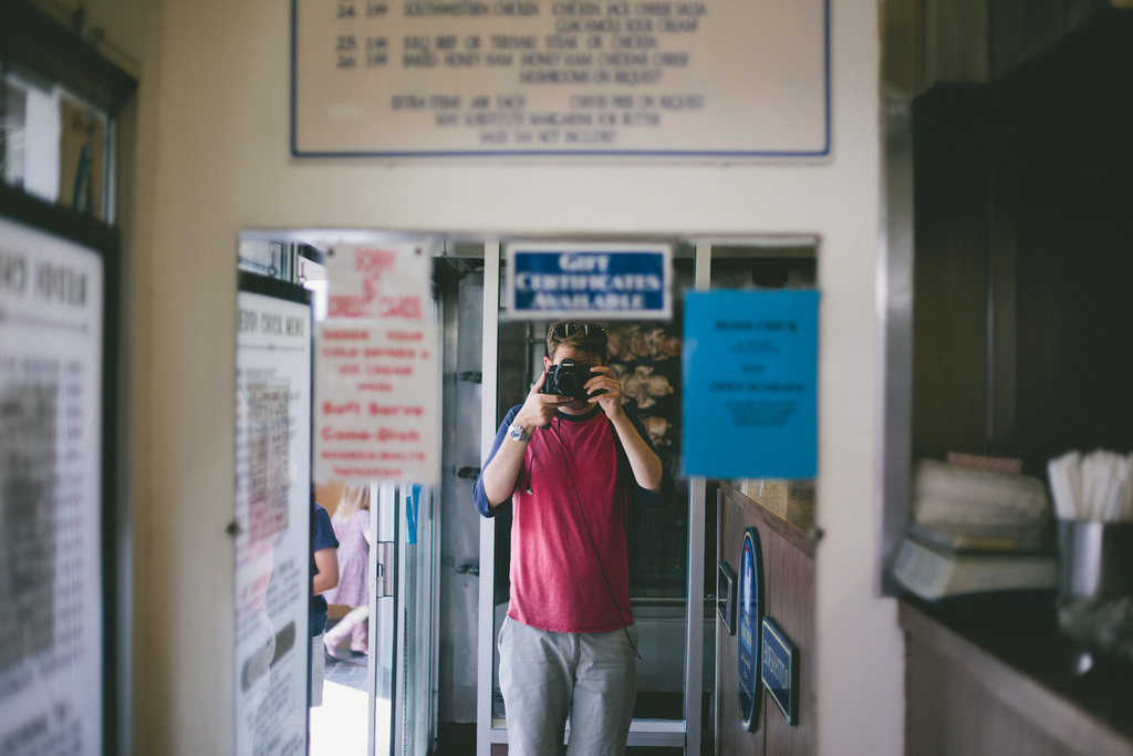 Mirror reflection of a photographer holding a camera in a retro diner surrounded by menus and signs.