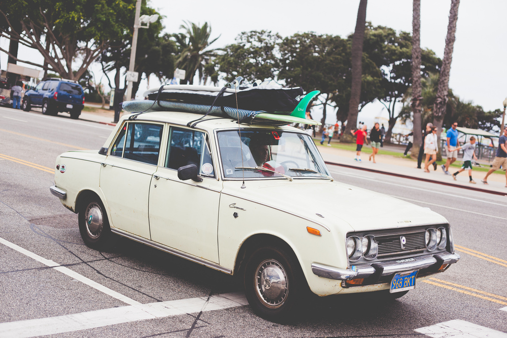 Vintage Toyota Corona sedan driving along a coastal California road with surfboards on its roof.
