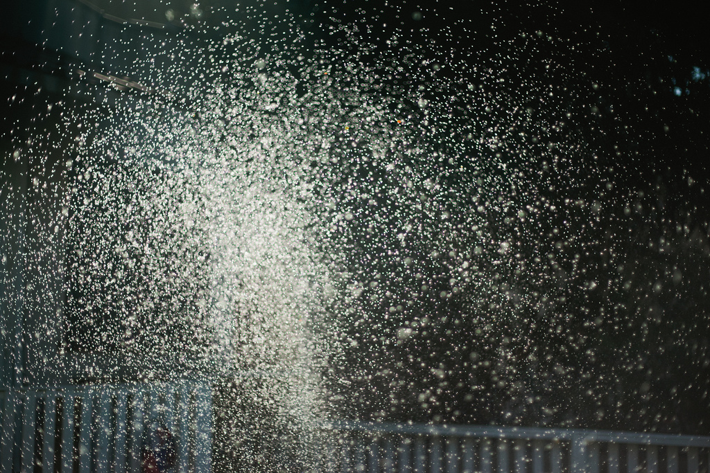 Backlit water droplets from an outdoor sprinkler catching late-day sunlight against a dark background.