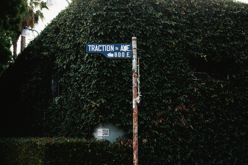 Street sign for 'TRACTION AVE. 800 E.' on a sticker-covered pole in front of an ivy-covered wall.