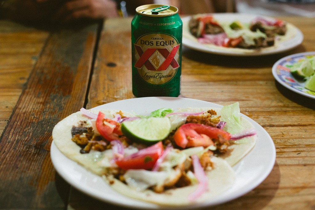 Plate of street tacos with tomatoes, onions, and lime next to a can of Dos Equis Lager Especial.