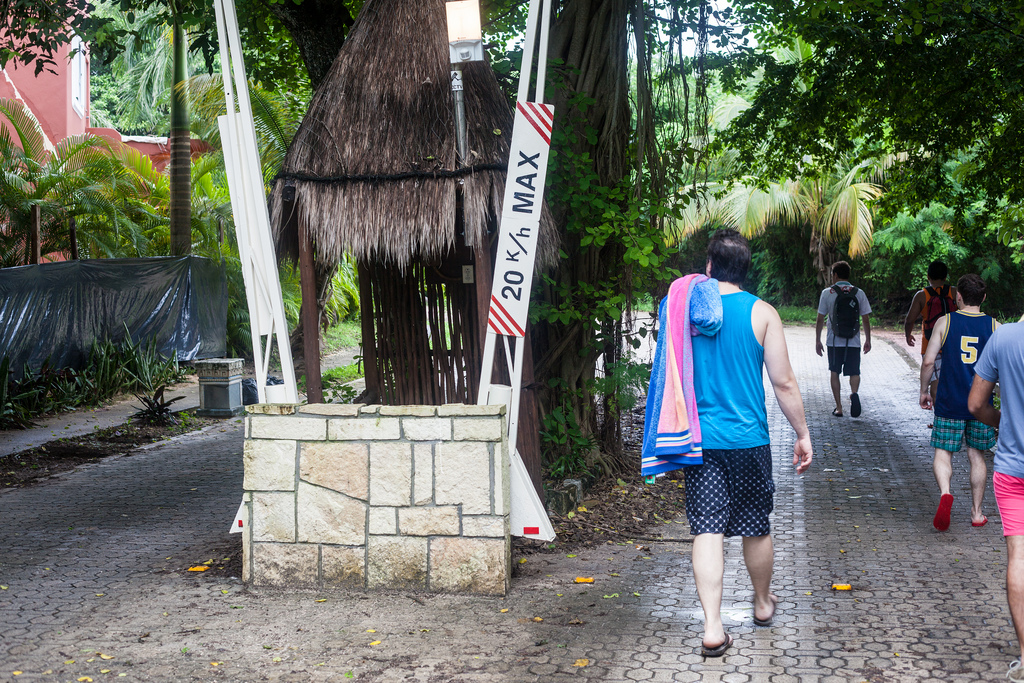 Friends walking past a thatched guard shack and a security gate arm marked '20 km/h MAX'.