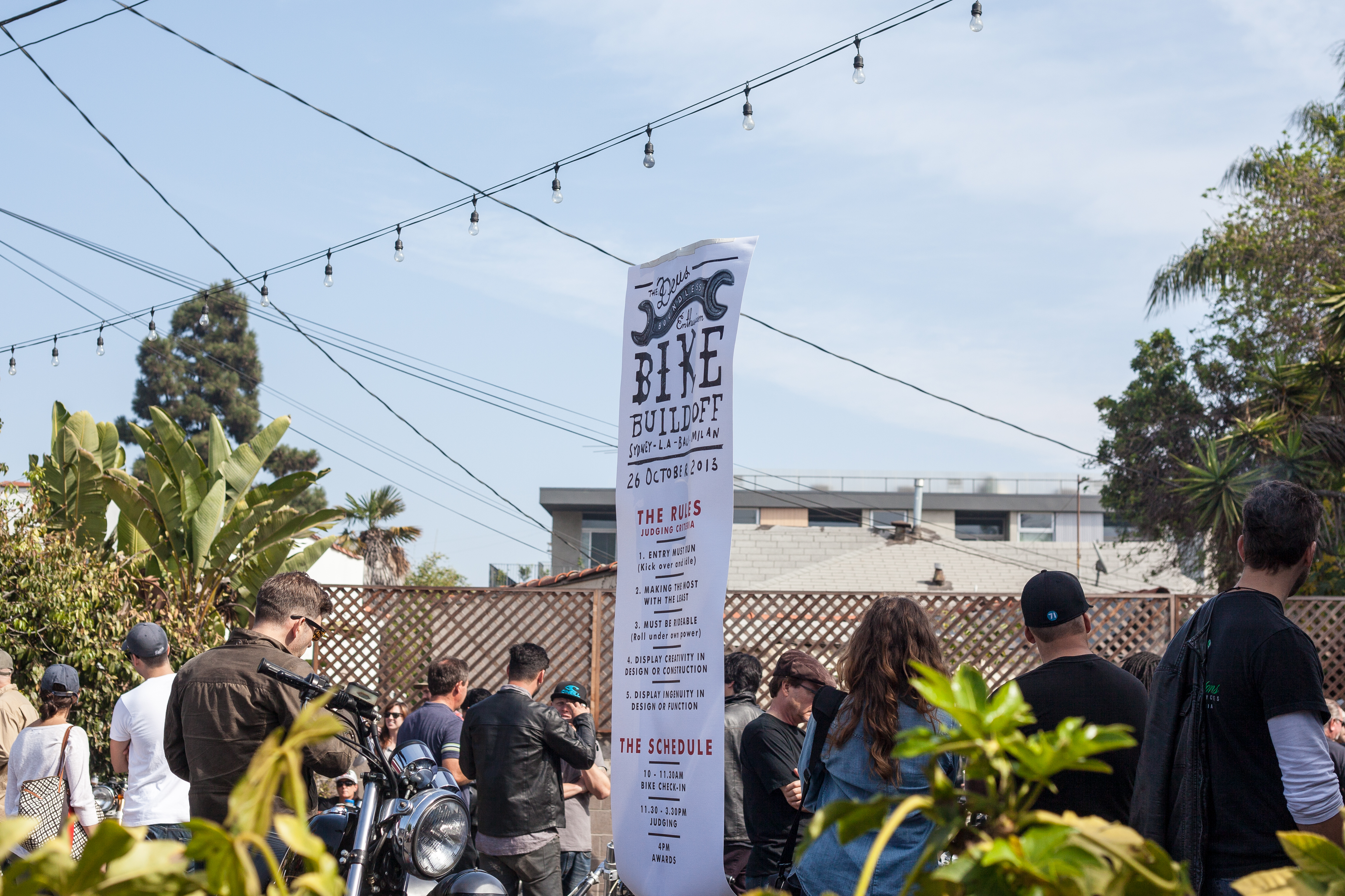 Banner for Deus Ex Machina Bike Build Off hanging above attendees and custom motorcycles.