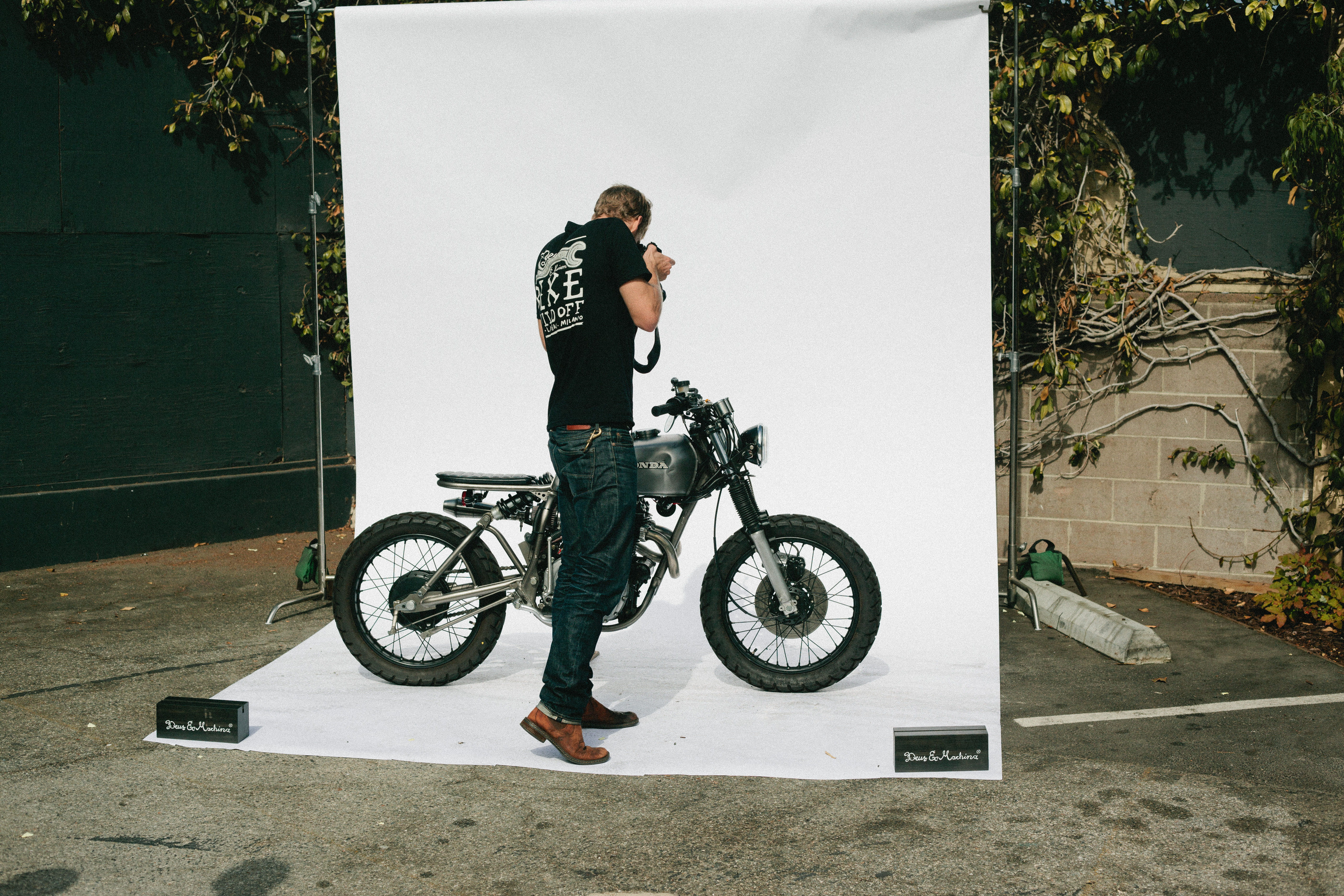 Photographer in a Deus shirt shooting a custom silver Honda motorcycle against an outdoor white backdrop.
