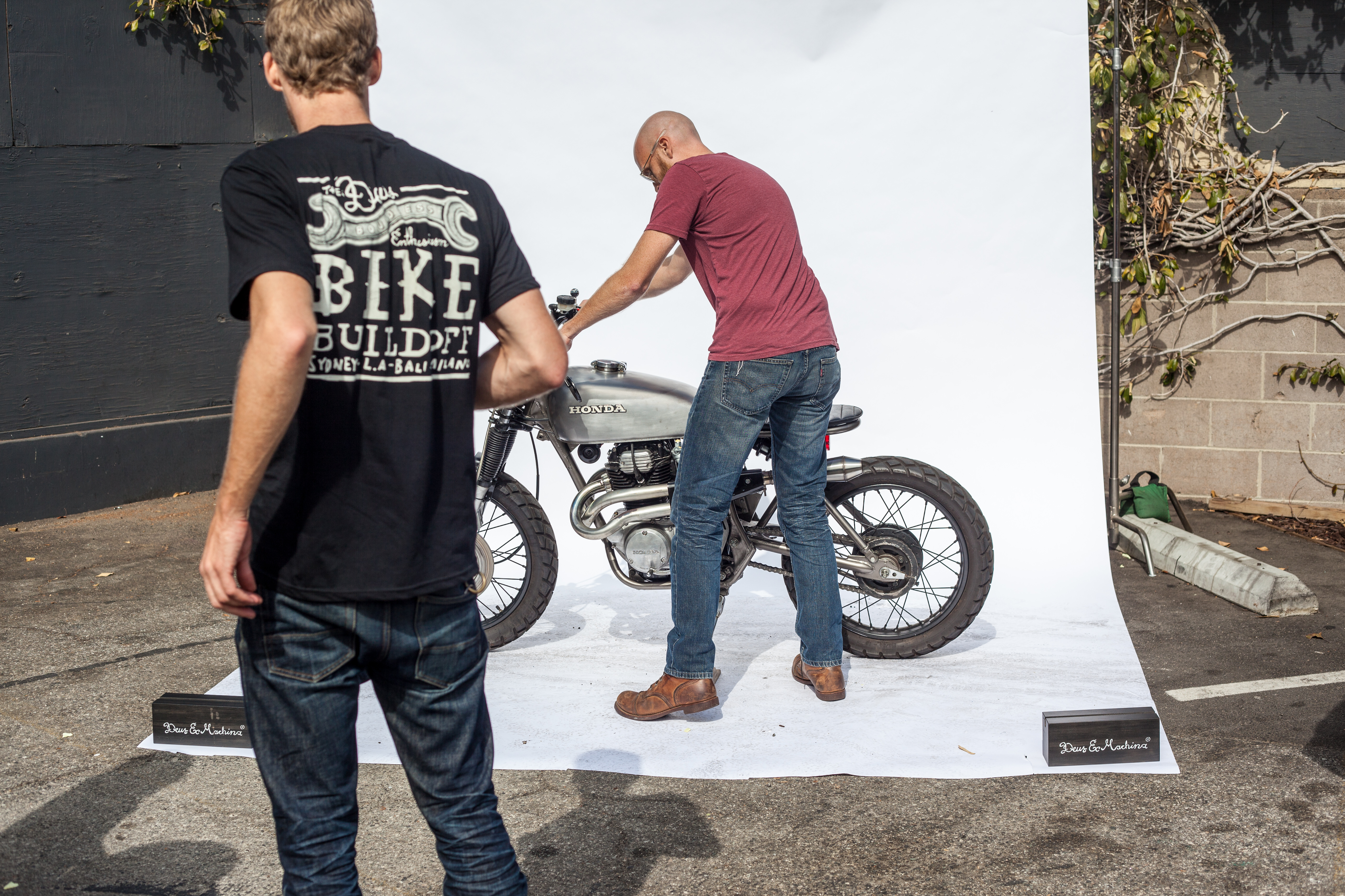 Man adjusting a custom silver Honda motorcycle on an outdoor white backdrop while a spectator watches.