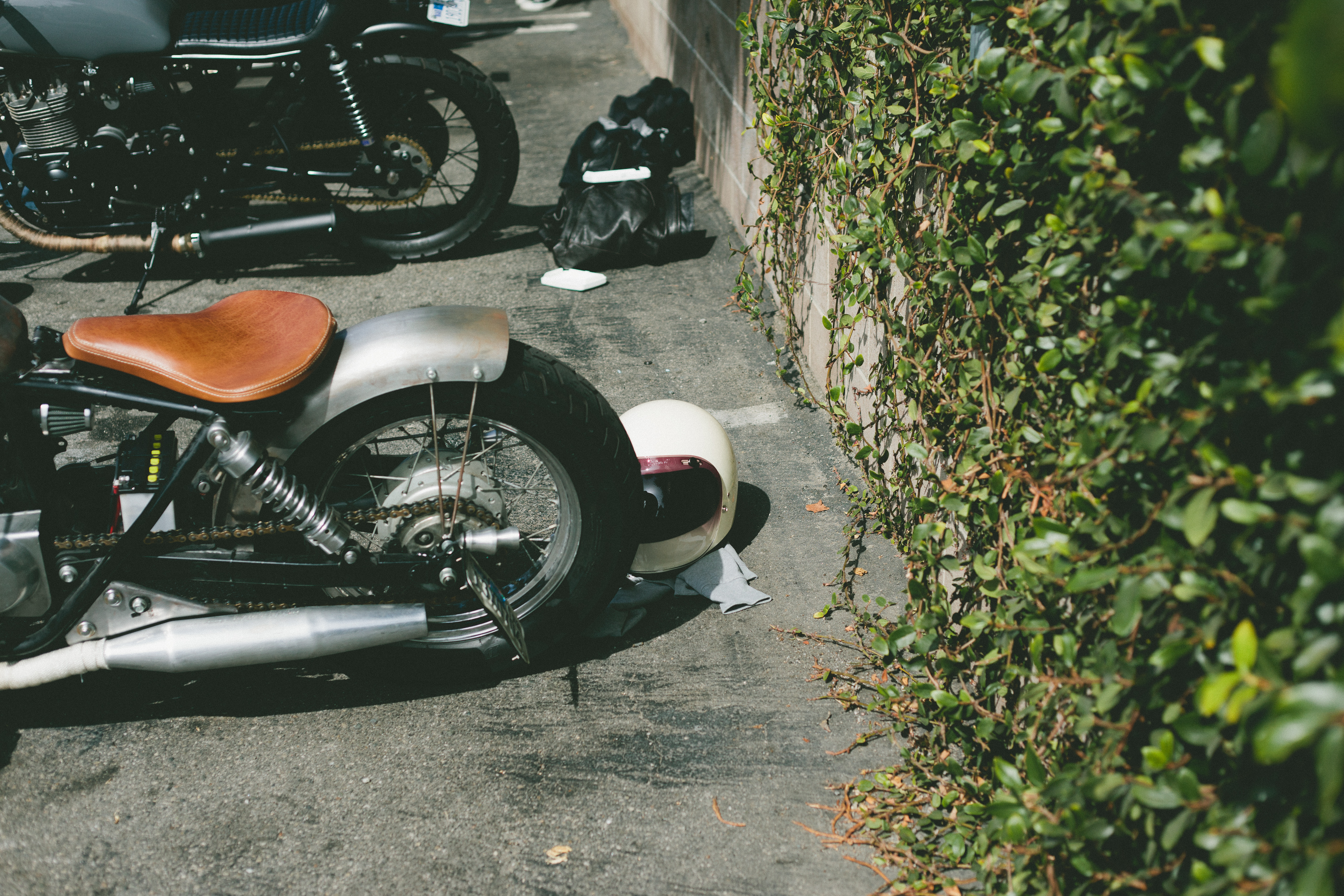 Custom motorcycle with brown leather saddle parked beside an ivy wall with a white helmet on the ground.