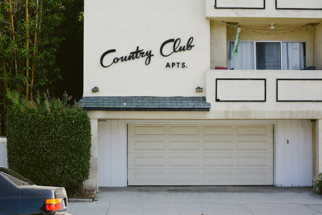 Exterior of a residential building featuring cursive black mid-century signage reading 'Country Club APTS.' above a white garage door and lush green foliage, shot with a Canon 5D Mark II.