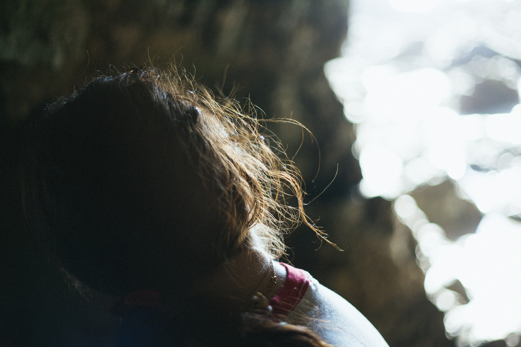 Close-up from behind of a woman's hair backlit by bright coastal sunlight pouring through a rocky sea cave opening.