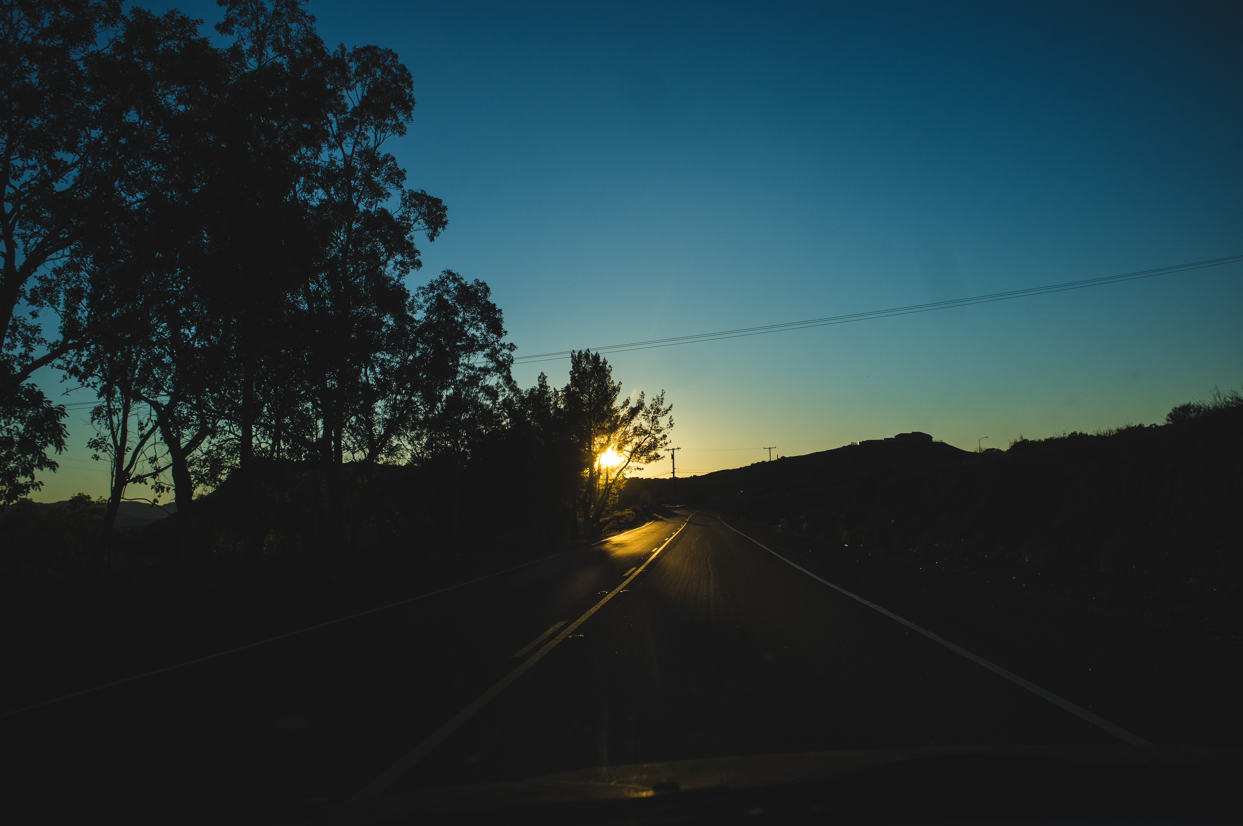 Driving along a two-lane country highway toward the setting sun bursting through roadside trees.