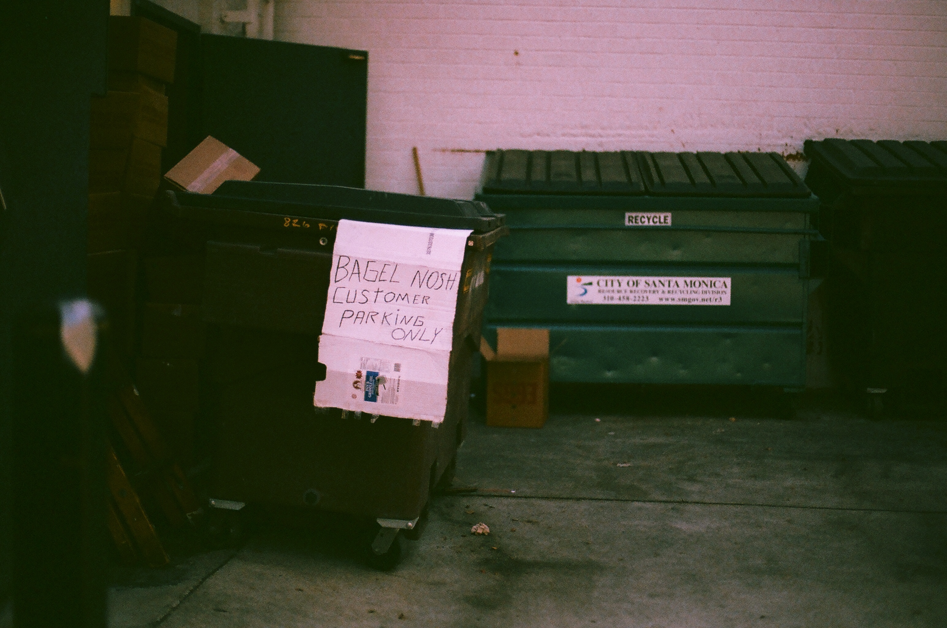 Dumpster sign reading 'BAGEL NOSH CUSTOMER PARKING ONLY' beside Santa Monica recycling bins.