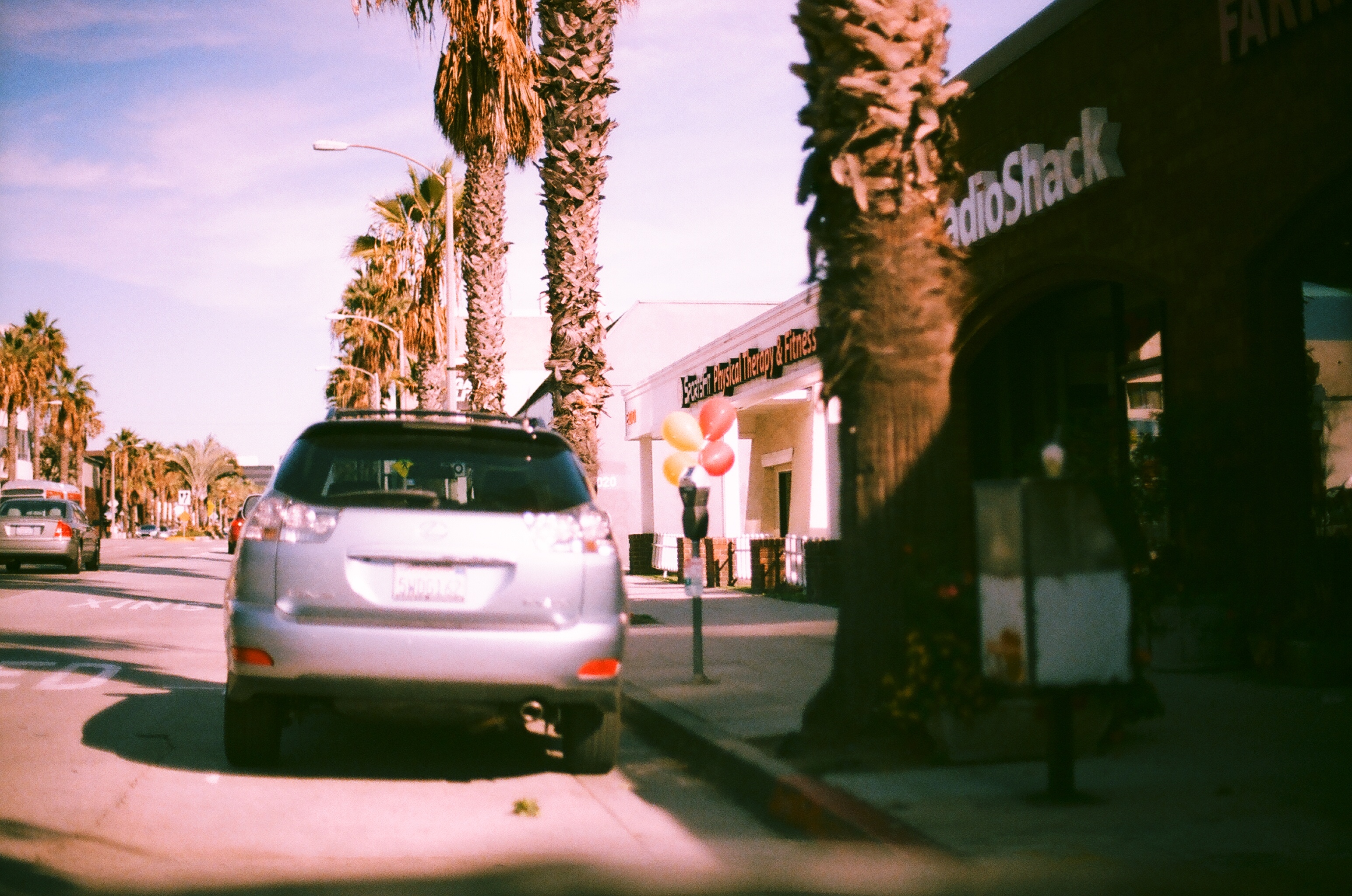 Silver SUV parked outside a RadioShack along a sunny, palm-lined street with balloons on a meter.