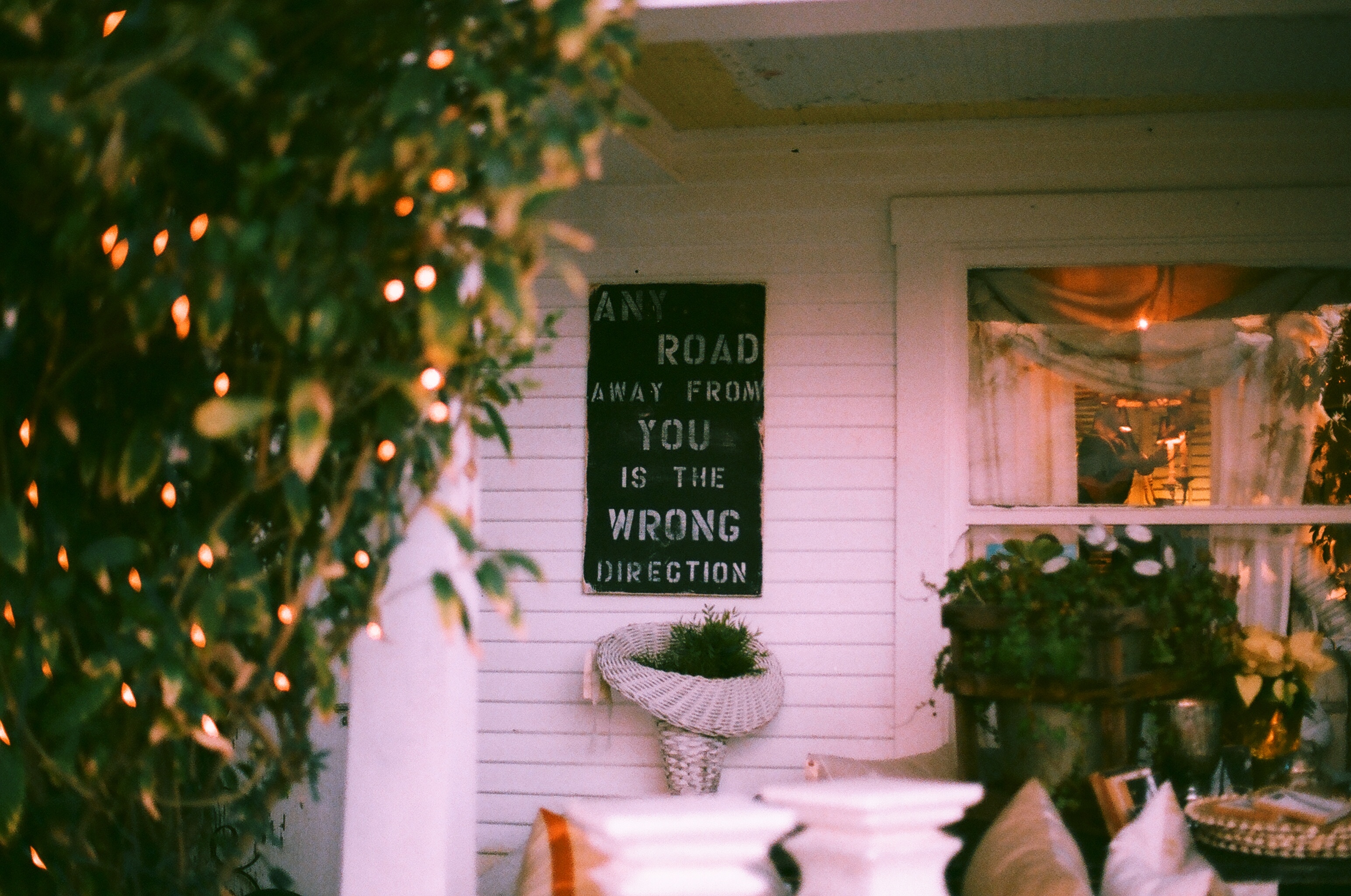 Sign reading 'ANY ROAD AWAY FROM YOU IS THE WRONG DIRECTION' on a white porch with fairy lights.