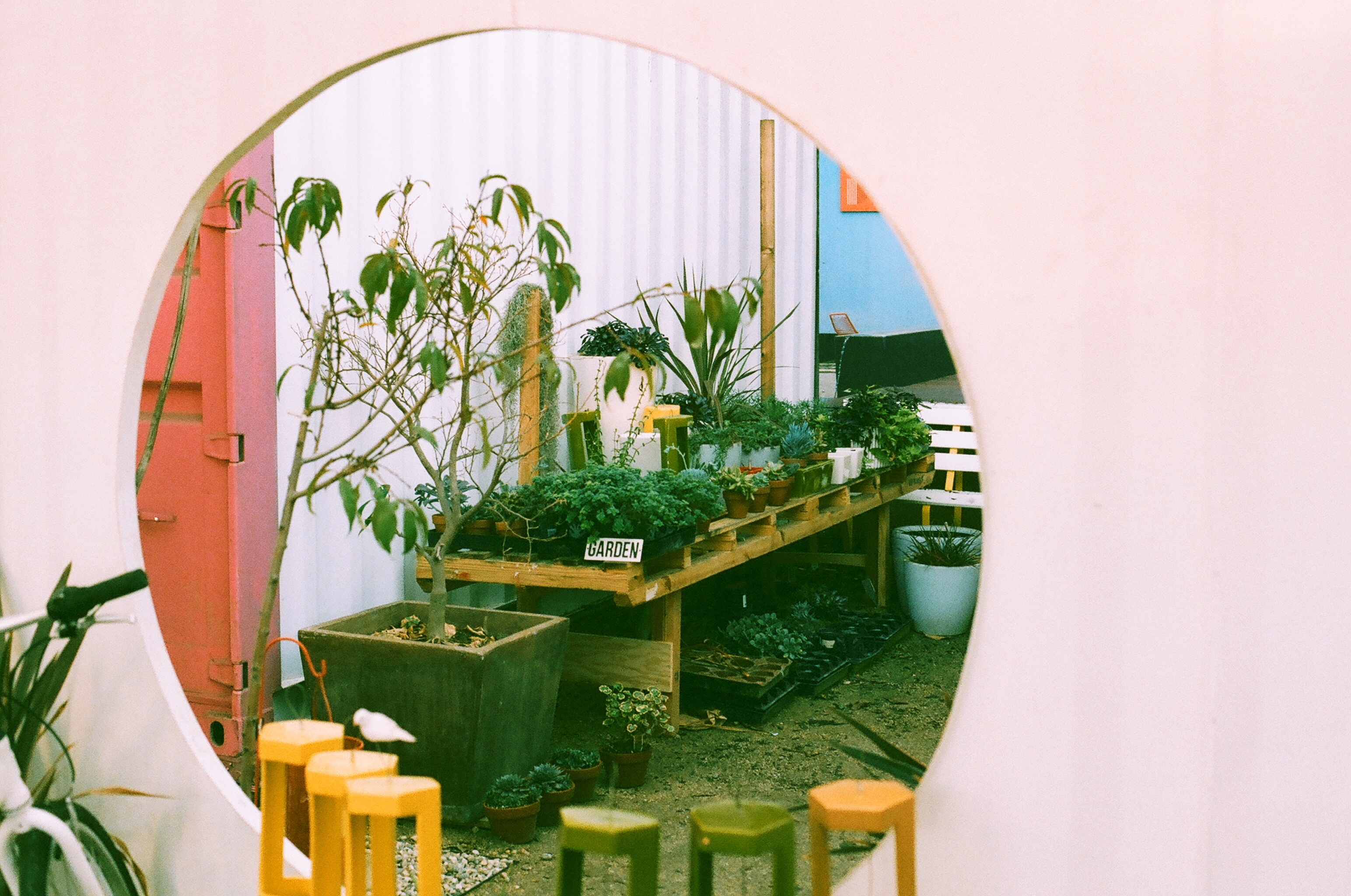 Circular wall mirror reflecting an outdoor plant nursery garden with a sign reading 'GARDEN'.