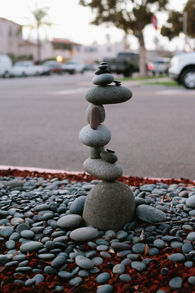 An intricate vertical rock cairn balanced delicately on top of smooth river stones in a garden bed with street traffic in the background, shot with a Canon 5D Mark II.