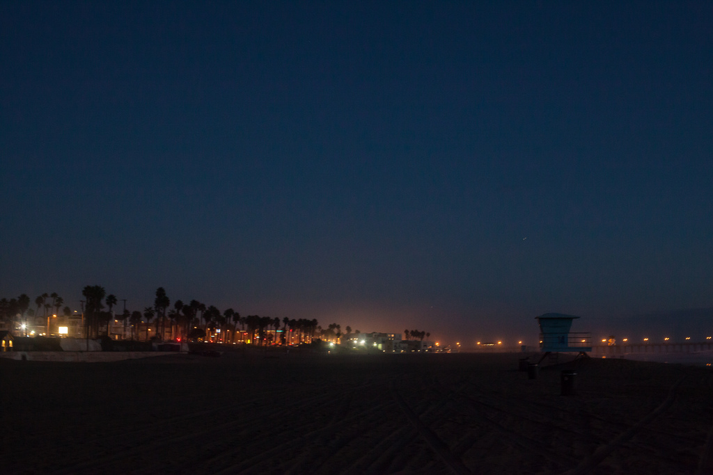 Nighttime view of Huntington Beach coastline looking across the dark sand toward a lifeguard tower and distant glowing city lights, shot with a Canon 5D Mark II.