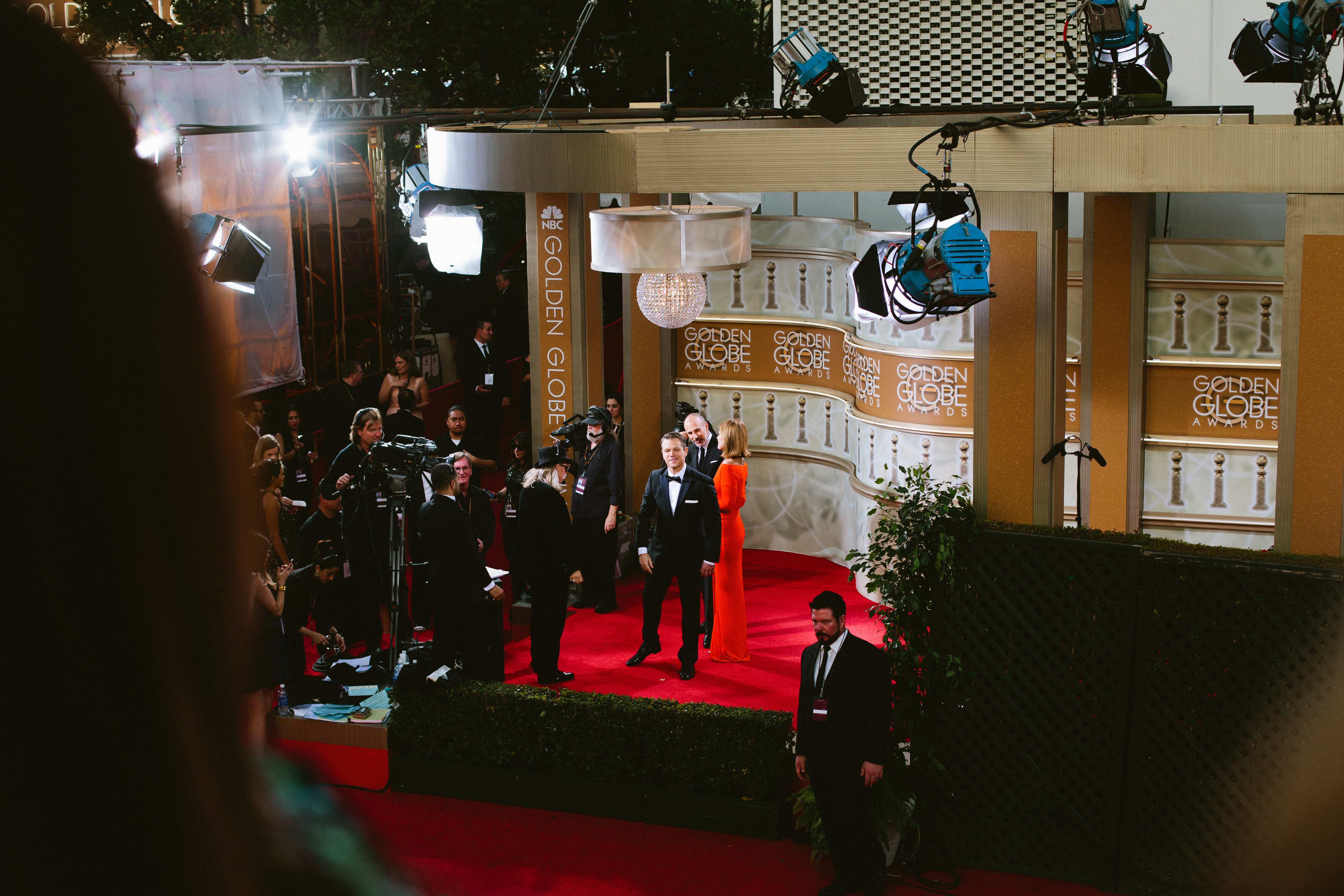 Matt Damon in a black tuxedo on the Golden Globes stage with a woman in an orange gown beside crew.