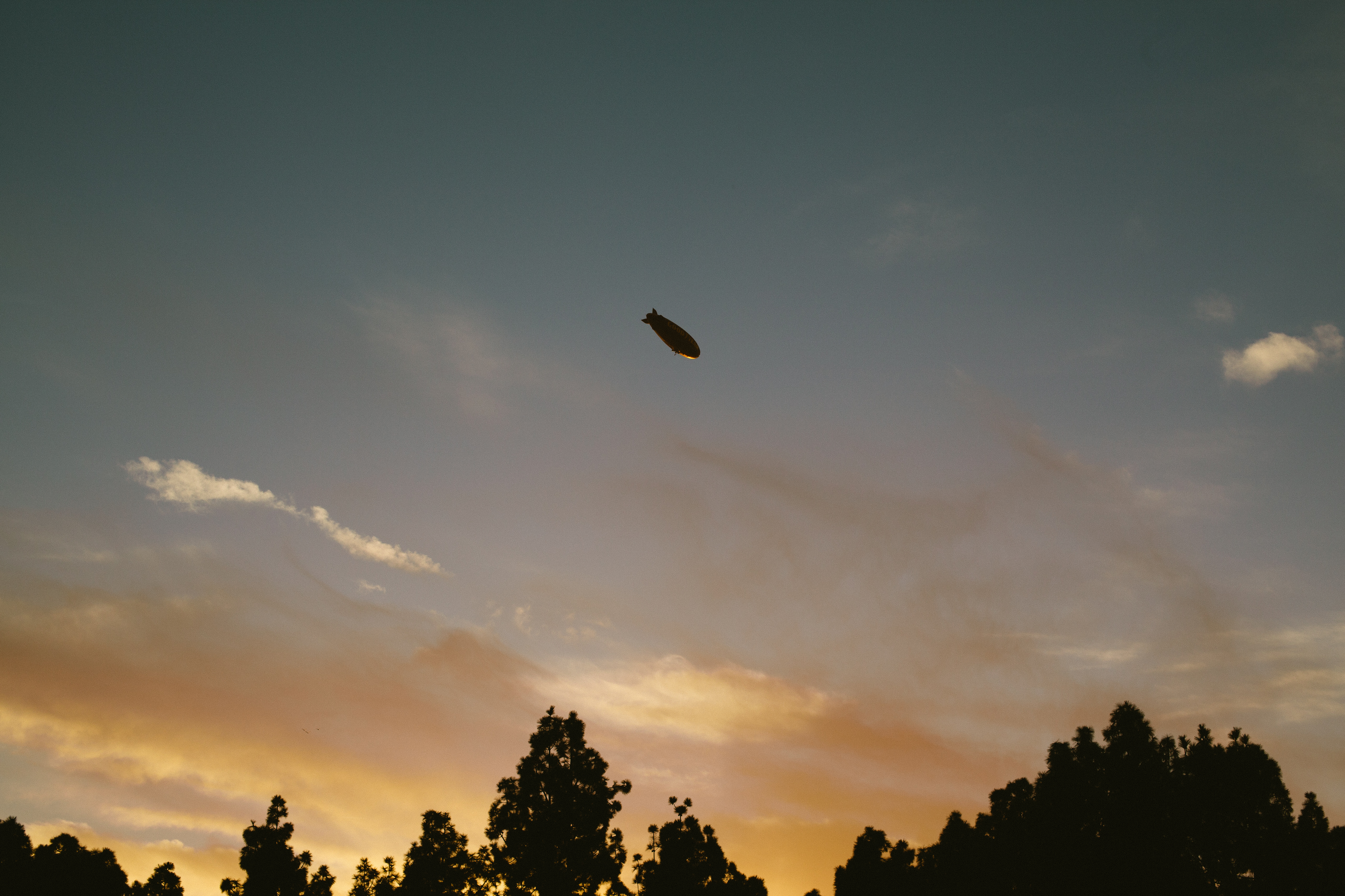 Goodyear blimp floating high in the dusk sky above tree silhouettes with soft orange and purple sunset clouds.