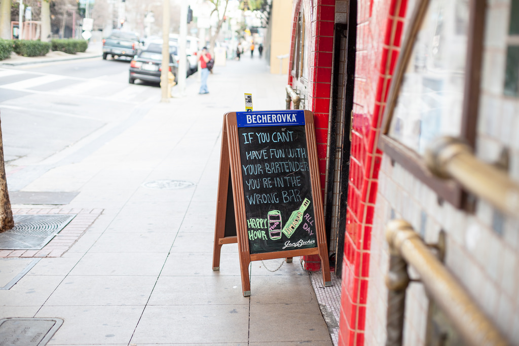 Sidewalk chalkboard sign outside a red-tiled bar reading 'IF YOU CAN'T HAVE FUN WITH YOUR BARTENDER YOU'RE IN THE WRONG BAR', shot with a Canon 5D Mark II.