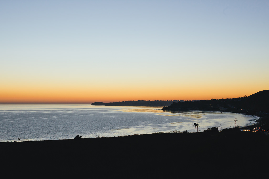 Panoramic sunset view overlooking the Malibu coastline and glowing Pacific waters beneath an orange and yellow gradient sky, shot with a Canon 5D Mark II.