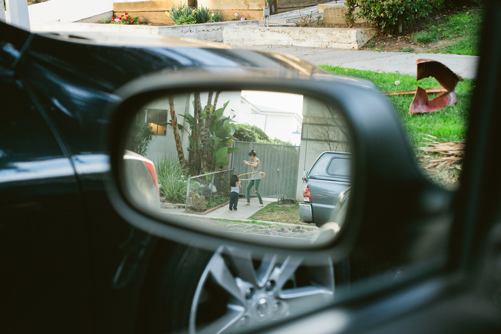 Side-view mirror reflection of a woman hula hooping on a sidewalk beside a child in a neighborhood.